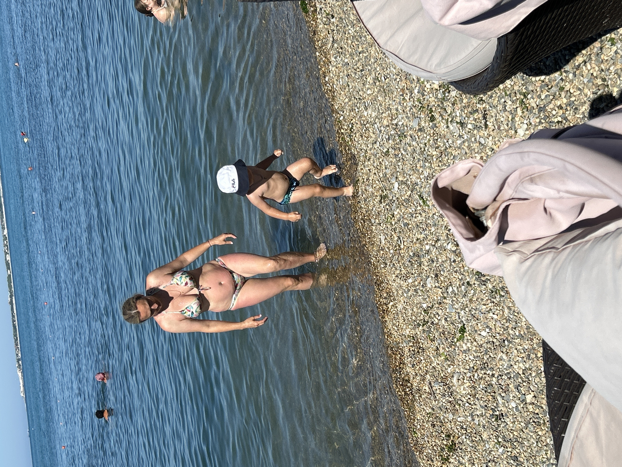 In Gelendzhik, Russia, on a sunny day, a woman wearing a floral bikini stands in shallow ocean water near a pebbled beach. A young child, wearing a white "FILA" hat and patterned swim trunks, walks out of the water towards the beach. In the background, other people are visible swimming in the sea, with a distant coastline and buildings suggesting a populated area. Two lounge chairs and a jacket are in the foreground, partially obscuring the view of the beach.