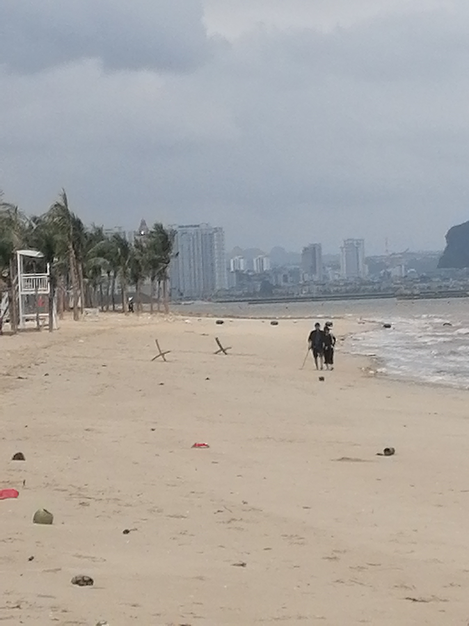 The image captures an overcast day at a sandy beach with a city skyline in the distance. The foreground features a wide expanse of light-colored sand, marked with numerous pieces of debris including discarded coconut shells and red plastic items. Two individuals, dressed in dark clothing, are visible walking along the waterline towards the right, with one holding a stick. The water shows small, gentle waves breaking on the shore.
To the left, a white wooden lifeguard stand or observation post stands prominently amidst a line of palm trees that stretch along the beach. Further into the midground, two wooden cross-shaped structures are stuck upright in the sand.
The background is dominated by a hazy cityscape, featuring multiple high-rise buildings under a cloudy sky. Distant mountains or hills are visible on the far right and behind the city, suggesting a coastal urban environment. The weather appears overcast with diffused light, indicating a cloudy day. No specific time of day is discernible, but it is clearly daylight. The scene suggests a public beach, possibly in an urban or semi-urban area, consistent with Bai Chay Ward in Vietnam. No discernible text is visible in the image.