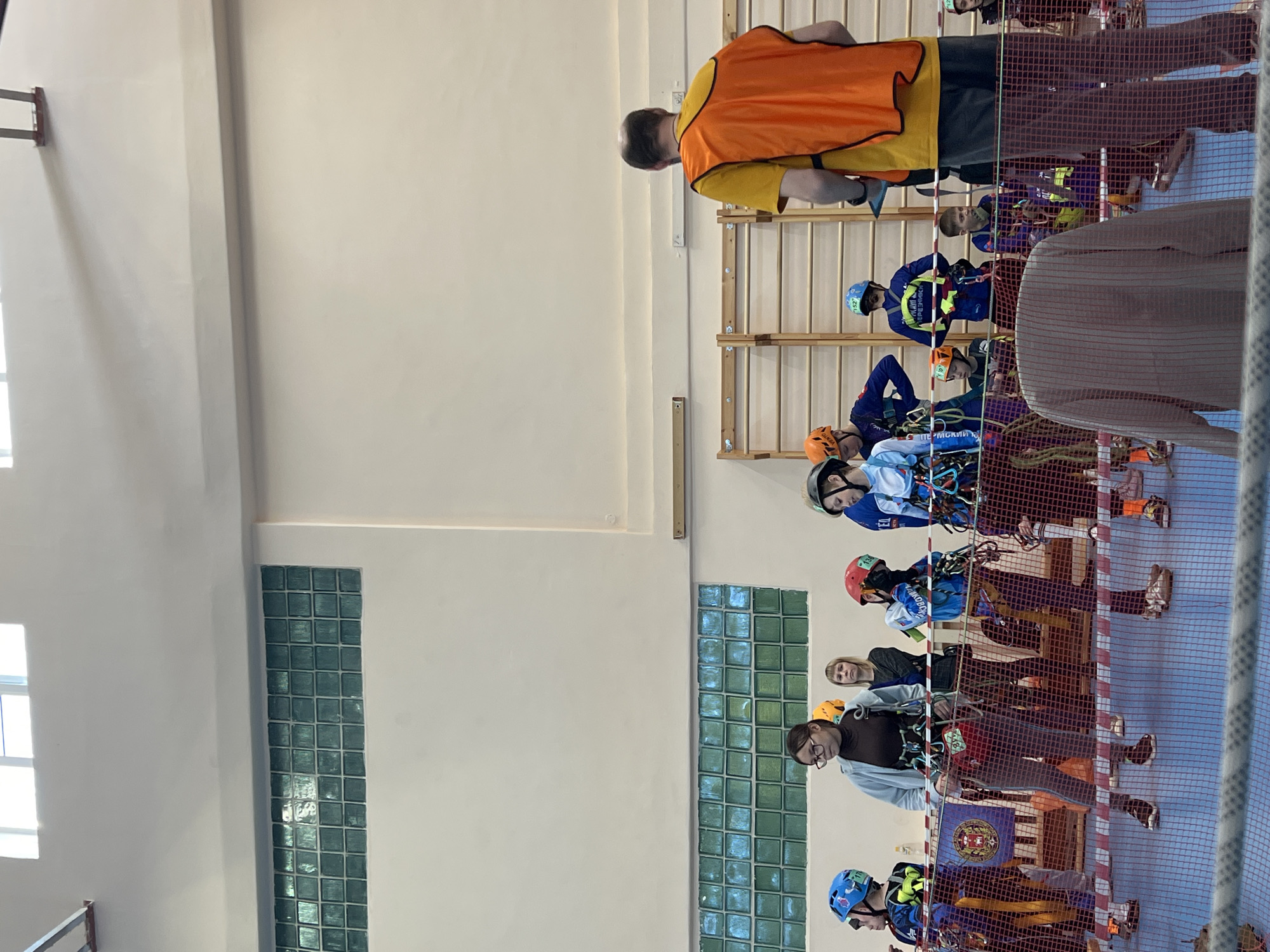 Location: Likely a gymnasium or indoor sports facility in Berezniki, Russia.
Scene: A group of people, mostly children, are wearing safety harnesses and helmets, suggesting they are participating in some kind of climbing or rope course activity. They are standing behind a net barrier. One adult male, wearing an orange vest, appears to be instructing or overseeing the group.
People:
* Several children wearing climbing harnesses and helmets
* At least one adult wearing an orange safety vest
* Another adult male with glasses standing with the group
* Possibly other adult supervisors/observers visible in the background
Subjects:
* Children preparing for a climbing activity
* Safety protocols and instructions
Objects:
* Climbing harnesses
* Helmets
* Rope
* Net barrier
* Climbing wall or equipment (partially visible)
* Wall bars (exercise equipment)
* Windows made of glass blocks
Actions:
* Children standing and listening
* Instructor providing guidance/supervision
* Possible preparation for climbing or rope course activity
Interactions:
* Instructor interacting with children
* Children interacting with each other
Environment:
* Indoor gymnasium or sports facility
* White walls
* Glass block window
* Bright lighting