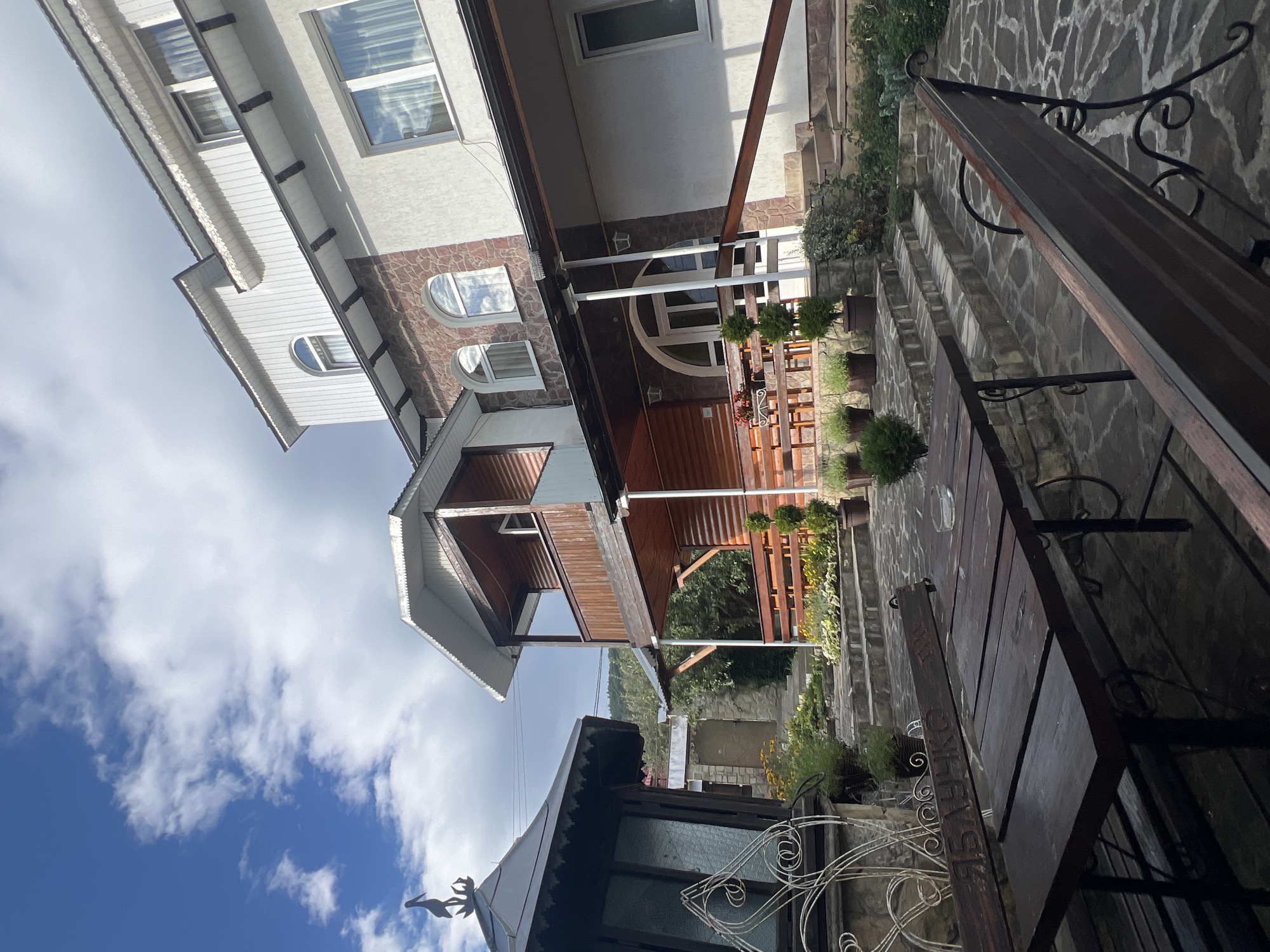The image captures an outdoor seating area adjacent to a multi-story building in Yaremche, Ukraine. The foreground features a dark wooden table with a simple metal frame and a glass ashtray on its surface, positioned on a stone-paved patio. A dark wooden bench with decorative metal legs is situated to the right of the table. Behind the seating area, a set of stone stairs ascends towards the building. The building itself is a mix of white stucco, stone, and wooden elements, with multiple windows and a covered balcony. To the left of the table, a dark wooden gazebo-like structure with decorative metalwork stands. Greenery, including potted plants and flowering bushes, is visible throughout the scene. The sky above is a mix of blue and white clouds. The setting appears to be a tranquil outdoor space designed for relaxation or dining.