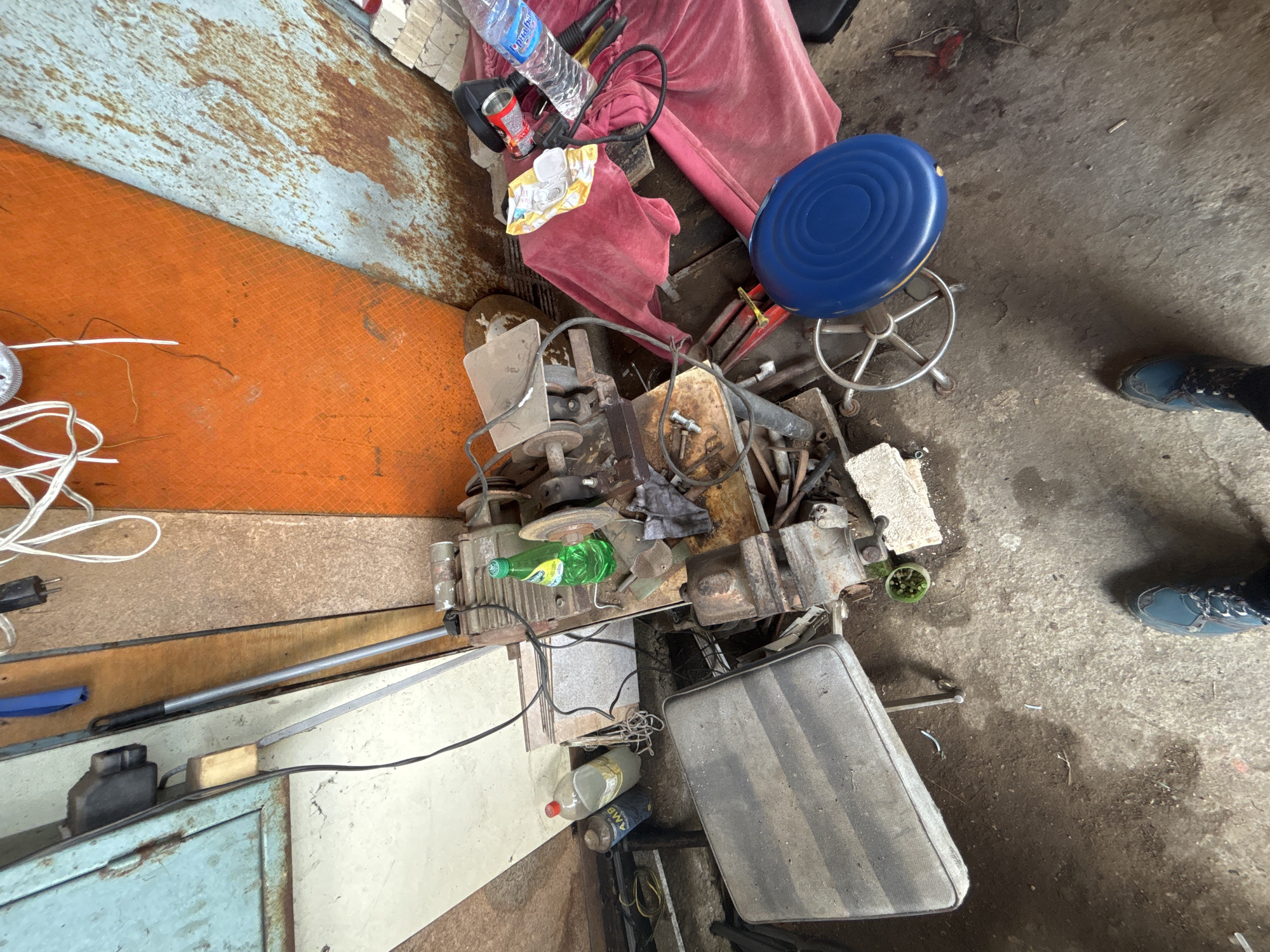 The image depicts a cluttered indoor space, possibly a workshop or storage area, in Zlatograd, Bulgaria. The perspective is a high-angle shot, revealing a variety of objects scattered across the floor and surfaces.
In the upper part of the image, there is a red fabric, a blue stool, and various discarded items including a water bottle and snack wrappers. Central to the image is a collection of mechanical components, possibly parts of an engine or machinery, along with various tools.
The flooring is a mix of concrete and wooden surfaces, showing signs of wear and disrepair. In the foreground, there's a dirty, upholstered chair and a cluttered floor.
The scene appears to be disorganized, suggesting a working environment that lacks regular cleaning and maintenance. The presence of tools and mechanical parts indicates that the space is used for repairs or manufacturing. A pair of shoes suggests that someone is present in the area or was recently there.