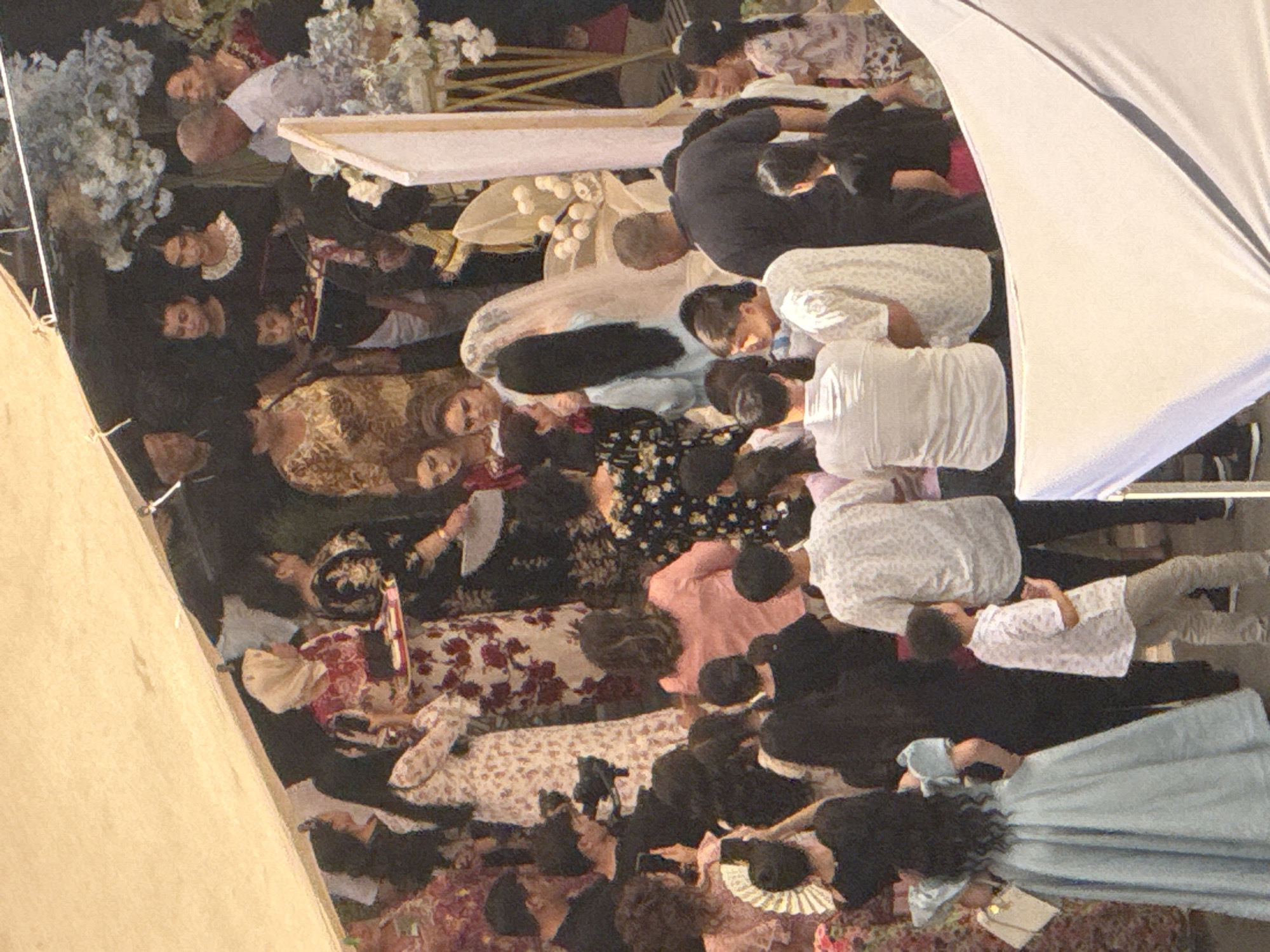 The image depicts a gathering of people, likely a wedding ceremony, taking place outdoors in Samarkand City, Uzbekistan. The scene is partially covered by a light-colored awning or tent.
Several individuals are visible, dressed in traditional and modern attire. A woman in a light blue, voluminous dress with puffed sleeves stands prominently in the foreground on the left, facing away from the camera. A white handbag is clipped to her left hip.
To her right, a group of men and women are assembled. Some are wearing headscarves. One woman in a white headscarf is holding a tray with what appear to be sweets or small items. Another woman is wearing a black dress with a floral pattern.
In the center and background, more attendees are present, including children. Some are observing the proceedings, while others appear to be engaged in conversation. A person in the far background, partially obscured, appears to be operating a video camera.
Floral arrangements, including white blossoms, are visible on the right side of the image, suggesting a celebratory event. The overall atmosphere appears festive and communal. The architecture and clothing styles suggest a cultural context specific to the region.