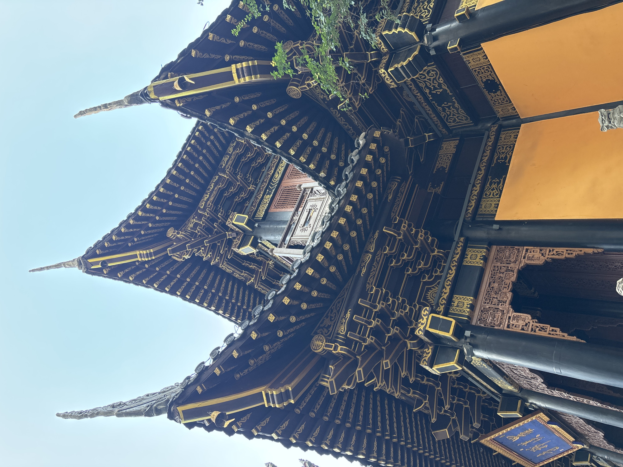 This media file captures an upward, angled view of the intricately decorated eaves and roof of a traditional Chinese building, likely a temple or palace, in Yuzhong District, China. The architecture is characterized by dark, wooden beams and panels adorned with elaborate golden patterns, including swirling motifs and repeated symbols. The rooflines are tiered and curved, with pointed finials extending outwards. Several layers of eaves are visible, creating a sense of depth and grandeur.
Against the dark wood and gold, sections of bright, golden-yellow panels stand out. Beneath the eaves, wooden beams and decorative elements showcase detailed carvings. A small blue plaque with golden figures, possibly depicting fish, is suspended from one of the beams. Green foliage from trees is visible peeking from behind one of the upper eaves, suggesting an outdoor setting, possibly with surrounding greenery. The sky is clear and light, indicating daytime, likely with bright sunlight given the strong contrast and illumination on the golden elements.
There are no people visible in the frame. The scene focuses entirely on the architectural details, emphasizing the craftsmanship and traditional aesthetic of the building. The overall impression is one of historical significance and cultural richness. No discernible text is present in the image other than the symbols incorporated into the decorative patterns.