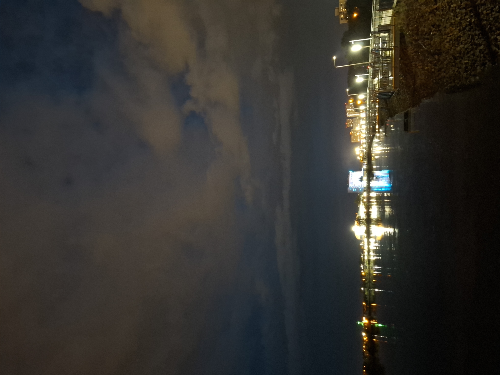 The image captures a nighttime view of the embankment along the Tura River in Tyumen, Russia. The sky is dark and cloudy, with some hints of orange and yellow from artificial lights. The river occupies the foreground and midground, reflecting the lights from the opposite bank and the embankment itself.
On the far bank, a city skyline is visible with numerous lights from buildings, some of which are illuminated and stand out against the darkness. There appears to be a larger, brightly lit building in the center of the cityscape, reflecting a vibrant blue light onto the water. Various smaller lights dot the shoreline, suggesting a developed urban area.
The embankment in the right midground and foreground features a walkway or promenade. Several lampposts with bright lights illuminate the area. There are outdoor seating areas, possibly for a cafe or restaurant, with tables and chairs visible. The ground along the embankment appears to be covered with gravel or small stones and some vegetation. The overall scene suggests a tranquil, urban nighttime setting by the river. No people are visible in the image.