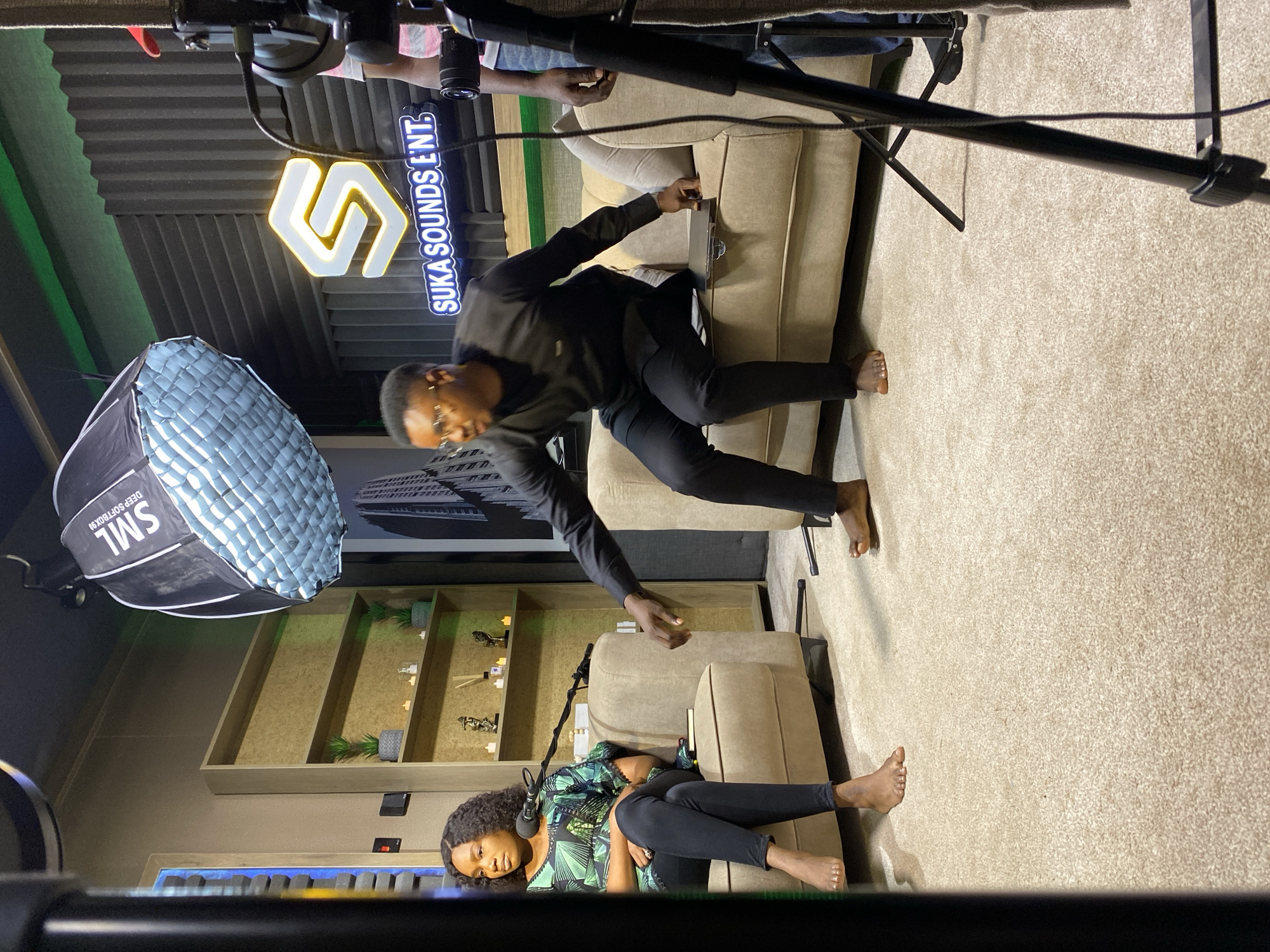 The image captures an indoor scene, likely a studio or living room, with a casual and relaxed atmosphere. Two people are present. In the foreground, a woman is seated on a beige sofa, wearing a green and black patterned top and black leggings. She has dark curly hair and is looking towards the camera. A microphone is positioned in front of her.
In the mid-ground, a man is standing and leaning over the same sofa, wearing a black top and black trousers. He is barefoot and appears to be interacting with the sofa or something on it. His pose suggests he might be reaching for something or adjusting it.
The setting includes a beige sofa and a carpeted floor. Behind the man, a wall with soundproofing foam is visible, along with a lit sign that reads "SUKA SOUNDS ENT". There's also a softbox light on the left side of the image. In the background, a shelf unit is visible with decorative items and plants.
The lighting in the scene appears to be artificial, suggesting an indoor setting possibly during daytime or nighttime, with the softbox indicating a professional setup for recording or photography. The overall mood seems informal and engaging, possibly during a recording session or a casual interview. The location context provided suggests the scene is in Bar Jirgi Summa, Nigeria.