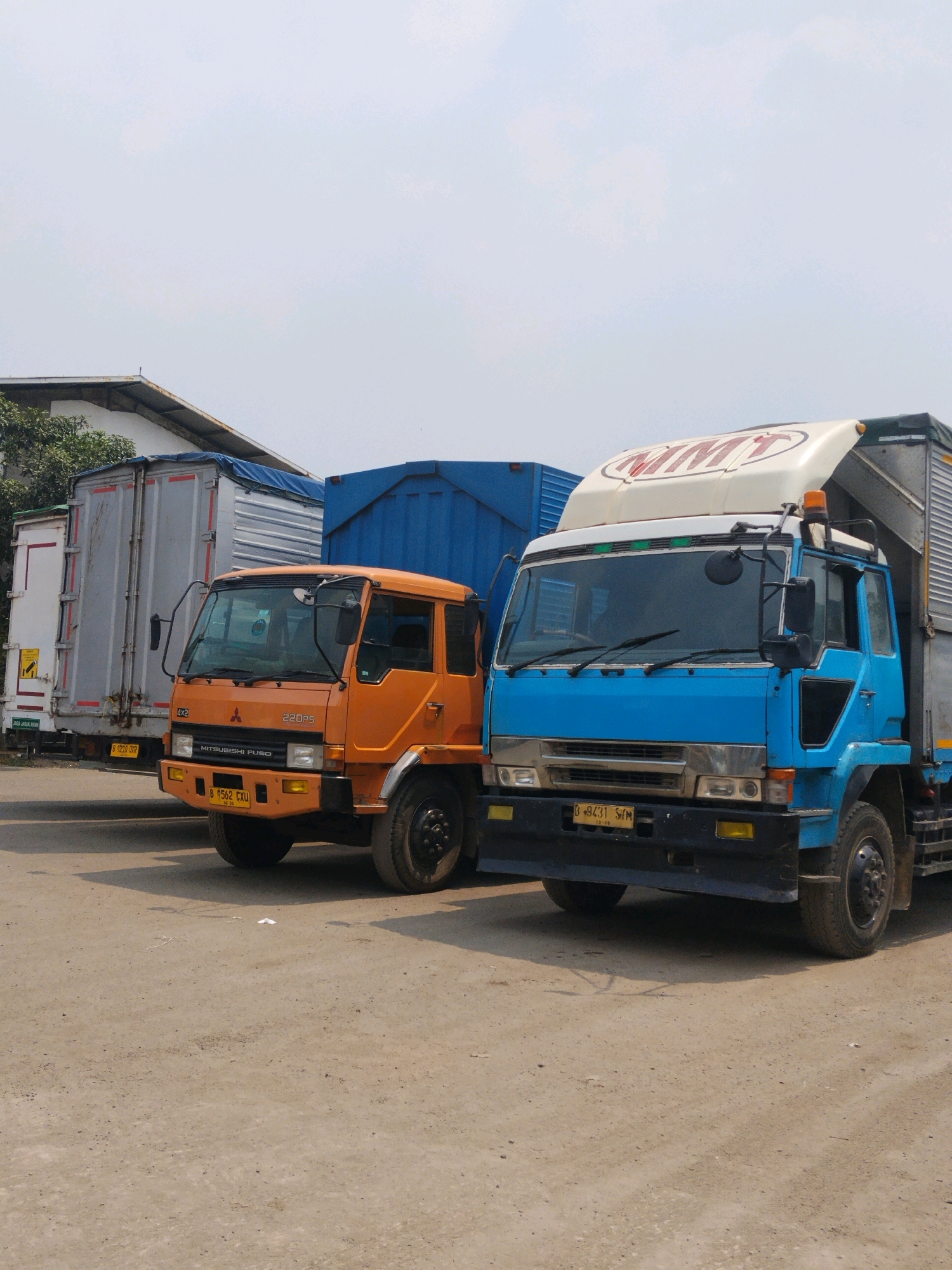 The image depicts a parking lot with several trucks. In the foreground, a bright blue truck is parked next to an orange truck. The blue truck has a white spoiler on top with red lettering that appears to say "MMT". The orange truck has "MITSUBISHI FUSO" written on its front. Behind these trucks, a silver-colored truck with a blue tarp is visible, and further back, a white truck with a yellow warning sticker is parked. The ground is a dirt or gravel surface, and the sky is overcast. The location is identified as Suka Nagara, Indonesia. There are no people visible in the image.