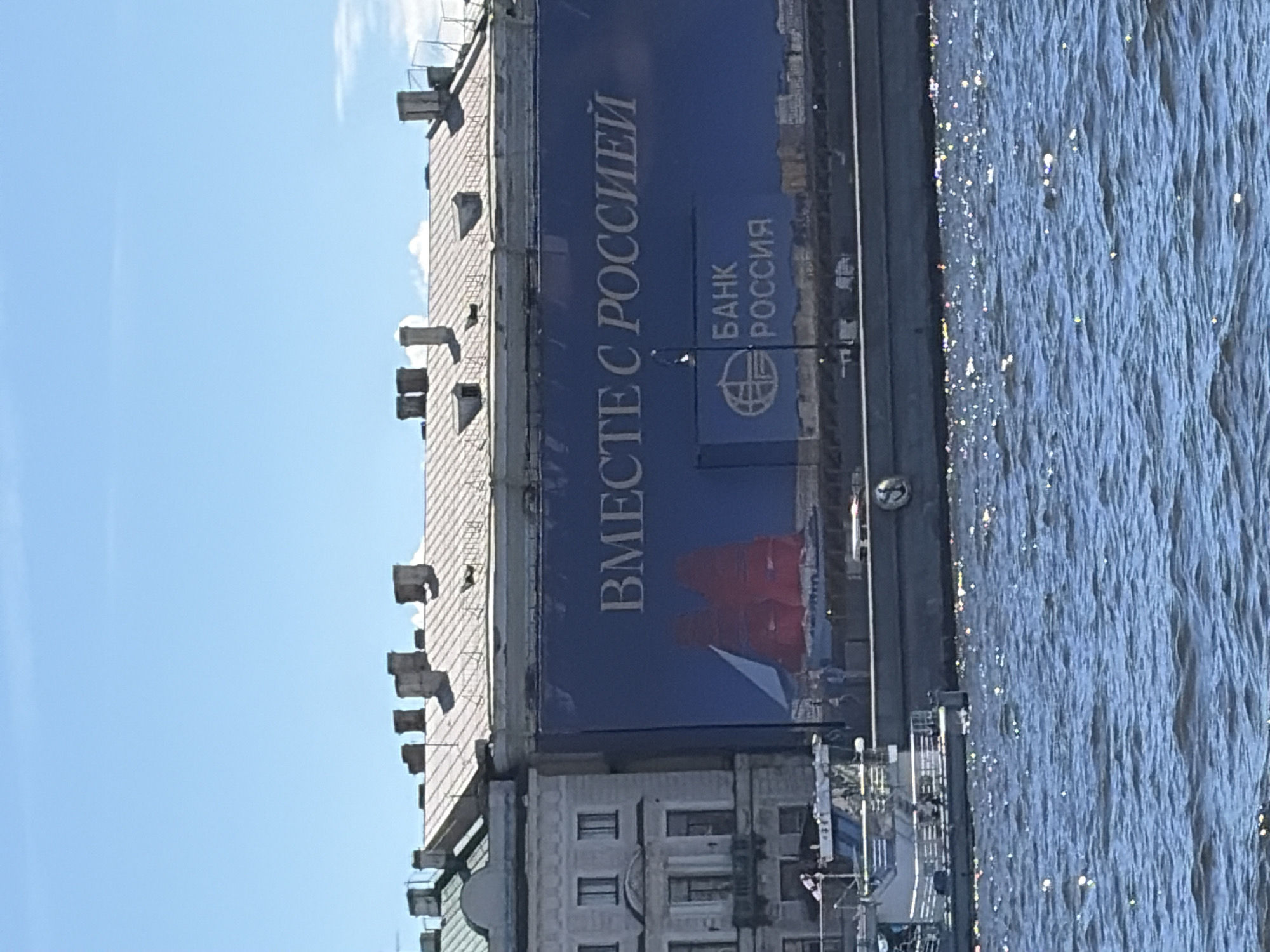 A wide, blue banner is draped across the side of a building, displaying text in white Cyrillic lettering that reads "ВМЕСТЕ С РОССИЕЙ" (Together with Russia). Below this, a smaller blue rectangular sign features the logo of "БАНК РОССИЯ" (Bank of Russia). In the foreground, a body of water, likely a river or canal, reflects the sunlight, creating a shimmering effect. A white and silver boat with multiple levels and railings is docked on the left side of the frame. The building itself is light-colored with multiple windows and a sloped roof topped with several chimneys. The sky above is a clear, bright blue with a few wispy white clouds visible. The overall scene suggests an urban waterfront setting in Russia.