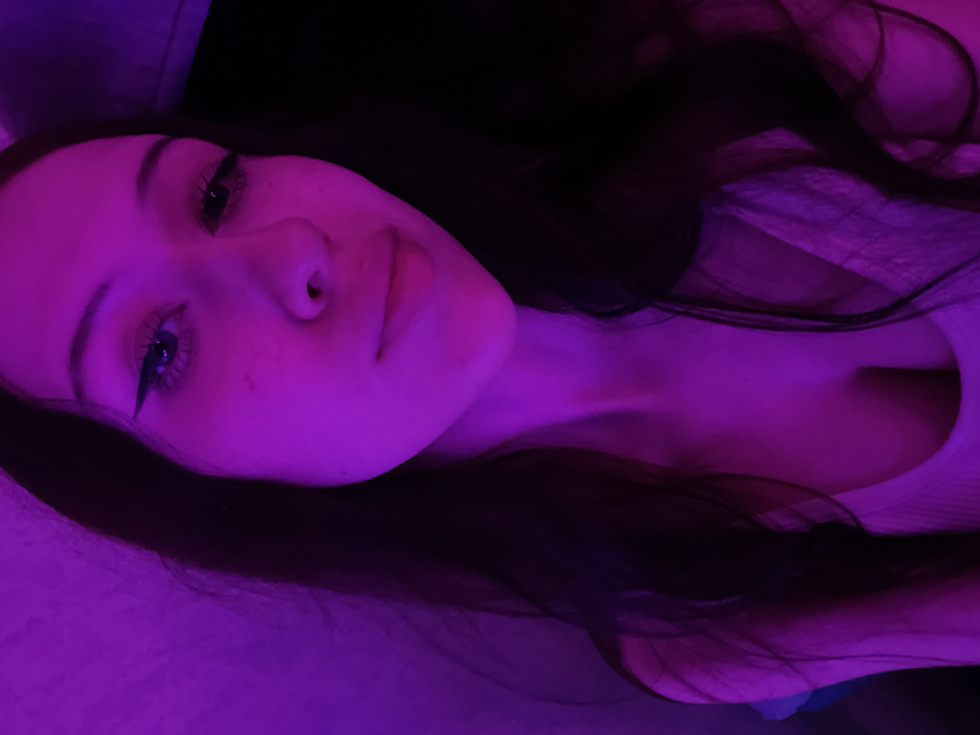 The image is a close-up, waist-up, selfie of a young woman with long, dark hair. She is illuminated by purple lighting, which casts a strong color cast over the scene and creates deep shadows. The woman is looking directly at the camera, with her eyes open and her lips slightly parted. She is wearing a light-colored top with a low neckline. The background is dark and out of focus, with a textured surface visible on the left side. There is no discernible location in Votkinsk, Russia, or any other specific geographical indicator within the image. The scene is a personal portrait captured in an indoor setting with artificial lighting.