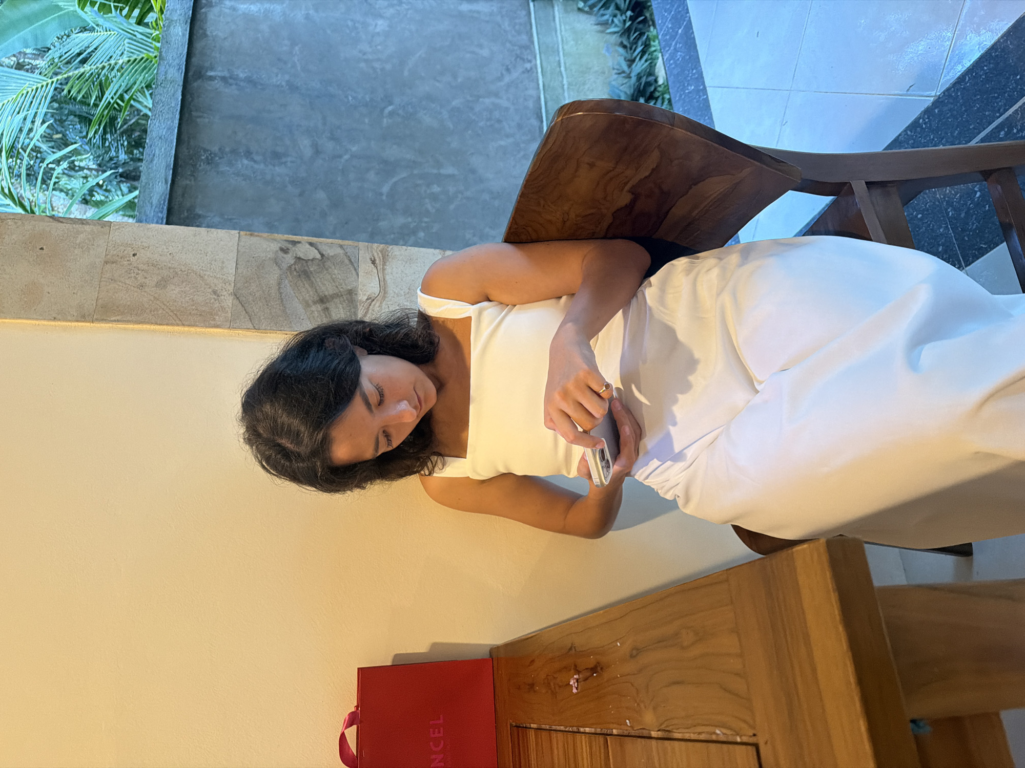 A woman is sitting in a wooden chair, holding a cell phone, and looking down at it. She is wearing a white tank top and white pants. Beside her is a wooden table and a red box with the word "CANCEL" printed on it. In the background, there is a window with plants visible outside. The location is Kedewatan, Indonesia.