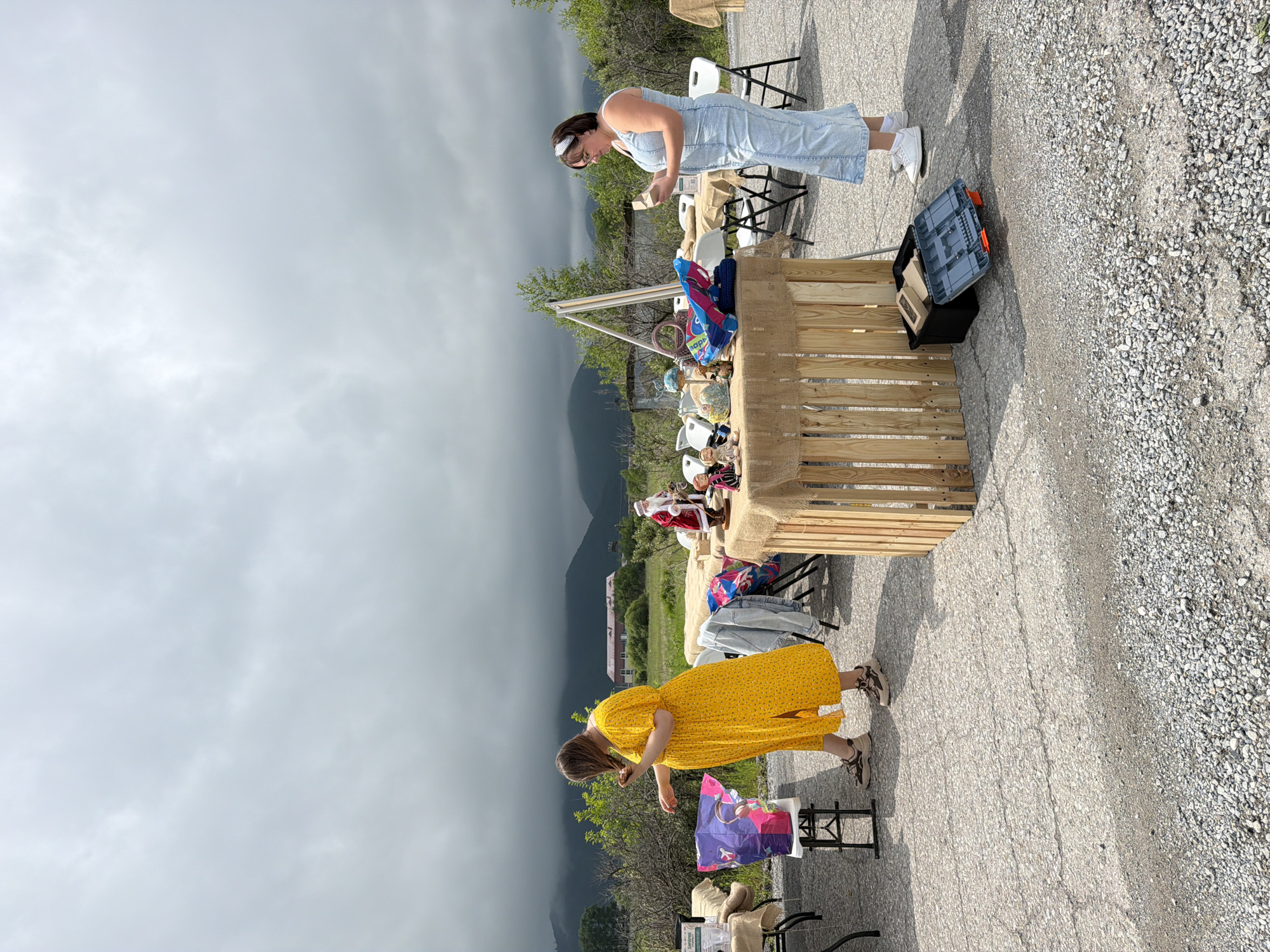 This image depicts an outdoor market or yard sale in Теплоозёрск, Russia, under a cloudy sky. Two women are present. The woman on the left, wearing a yellow polka-dot dress and sneakers, is reaching into a colorful bag on a folding table. She has brown hair. The woman on the right, in a light blue denim dress and white sneakers, is holding a small object and looking down at it. She has dark hair and wears glasses and a white headband. Several tables draped with burlap are set up, displaying various items including a Santa Claus figurine, dolls, bags, and a wooden crate containing a toolbox and other objects. In the background, mountains are visible, with some buildings and trees in the middle ground. The ground is paved with asphalt and gravel.