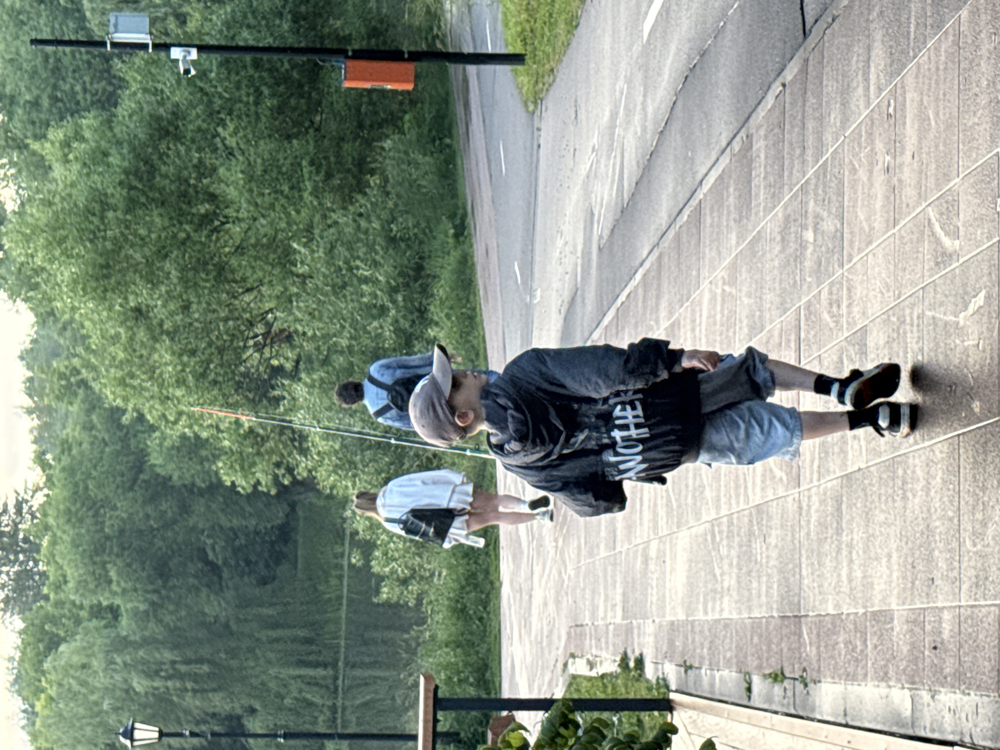 In Moscow, Russia, a young boy walks away from the camera on a paved walkway. He wears a dark hooded jacket with white lettering, light blue shorts, and black and white sneakers. He carries a fishing rod. Behind him, two people walk away from the camera on the same walkway. The person closer to the camera wears a white jacket and a backpack, and the person further away wears a light blue denim jacket and a backpack. The walkway is bordered by a body of water on the left and a road on the right, with dense green trees in the background. A lamppost is visible on the left side of the walkway, and a pole with an orange box and what appears to be a security camera is on the right side of the road. The sky is overcast, and the light suggests it is either early morning or late evening.