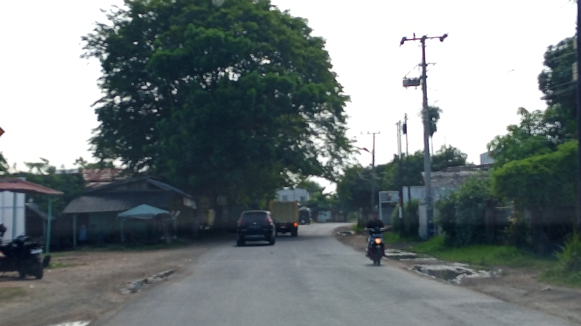The image depicts a street scene in Uleereuleung, Indonesia, with a focus on a paved road winding through a rural or semi-rural environment. On the left side of the road, there are several structures, including a building with a red roof and a darker structure with a covered area. A motorcycle is parked to the left, facing away from the camera. Further down the road on the left, there is a dark-colored SUV.
In the center of the road, a light-colored truck with a cargo bed is traveling away from the camera. To the right of the truck, another vehicle is visible. On the right side of the road, a person on a motorcycle is traveling towards the camera. The environment is characterized by lush green trees lining the road, with a bright sky overhead. Several utility poles are visible along the roadside, indicating the presence of electricity infrastructure. The road surface appears somewhat worn, with patches of dry earth and some puddles of water. The overall impression is that of a typical Indonesian street in a developing area, with a mix of vehicles and local architecture under a hazy sky.