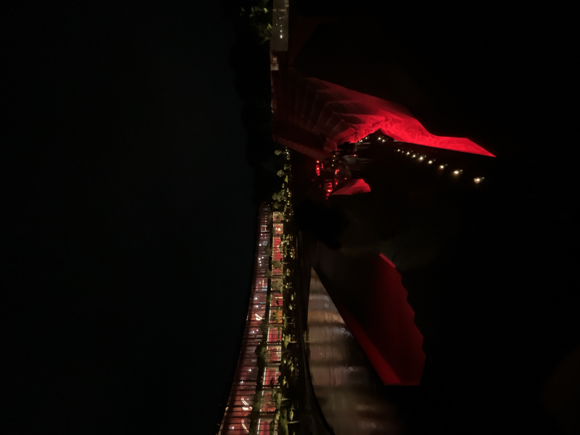 The image depicts a nighttime scene outside a modern, curved building with extensive red-tinted glass windows. The building is illuminated from within, casting a red glow. In front of the building, there is a body of water reflecting the light. A large, flowing red fabric installation is prominently featured, extending from the foreground and wrapping around a set of stairs or ramp-like structure. Small, warm lights are visible along the path of the red fabric installation, and a few silhouetted figures can be seen near the structure, suggesting an event or gathering. The surrounding environment is dark, with some trees and landscaping visible in the middle ground. The overall impression is one of an illuminated outdoor architectural display or event. The image is not verifiable to be located in Krasnodar, Russia.