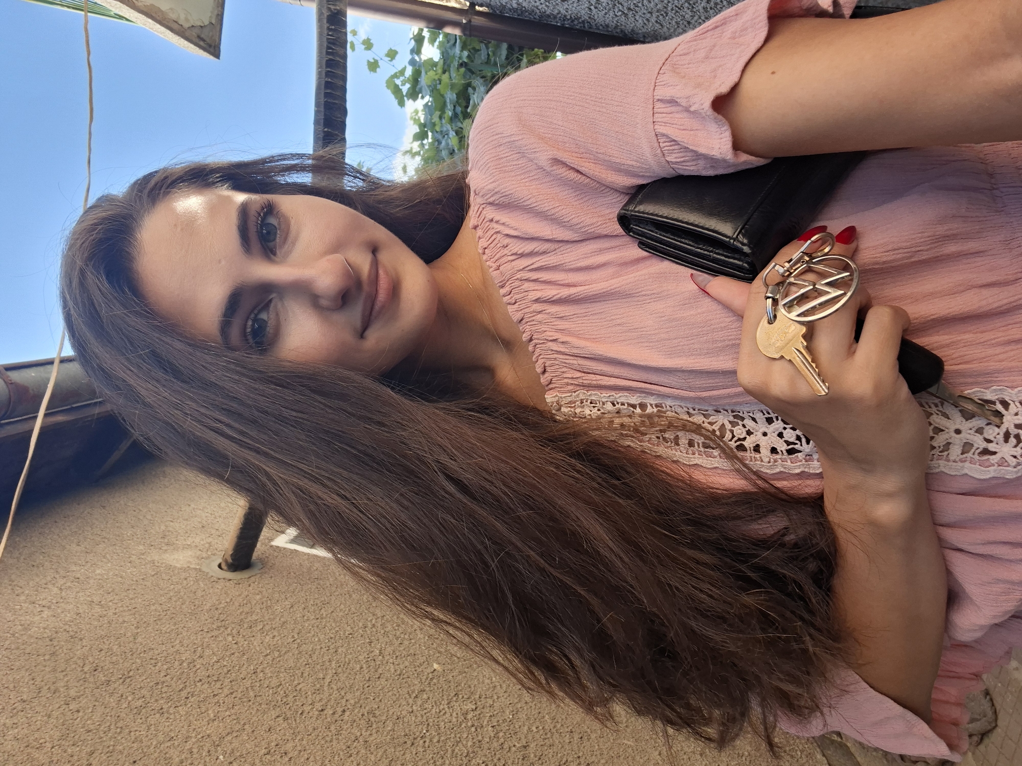 A young woman with long, dark hair is holding keys with a Volkswagen logo and a black wallet in her right hand. She is wearing a pink, ruffled blouse. The background shows a textured wall, a pipe, and a bright blue sky with some greenery. The scene appears to be outdoors, possibly in a residential area.