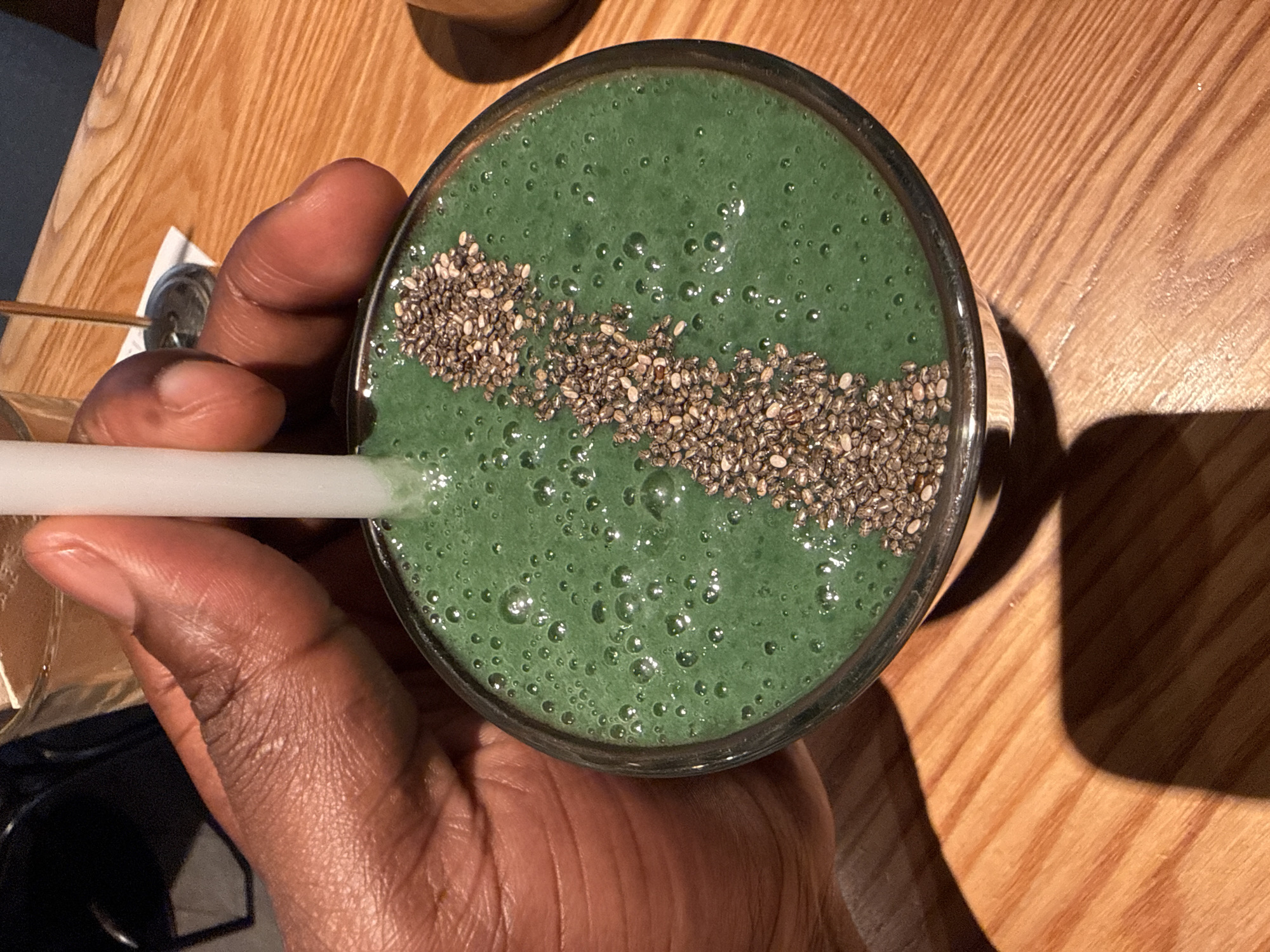 A close-up, high-angle shot shows a hand holding a clear glass of a green smoothie topped with a line of seeds, possibly chia seeds. A white straw is visible in the drink. The hand holding the glass appears to be dark-skinned. The drink sits on a wooden surface, presumably a table. The lighting suggests an indoor setting. No elements indicate a specific location or city in Yuzhong District, China.