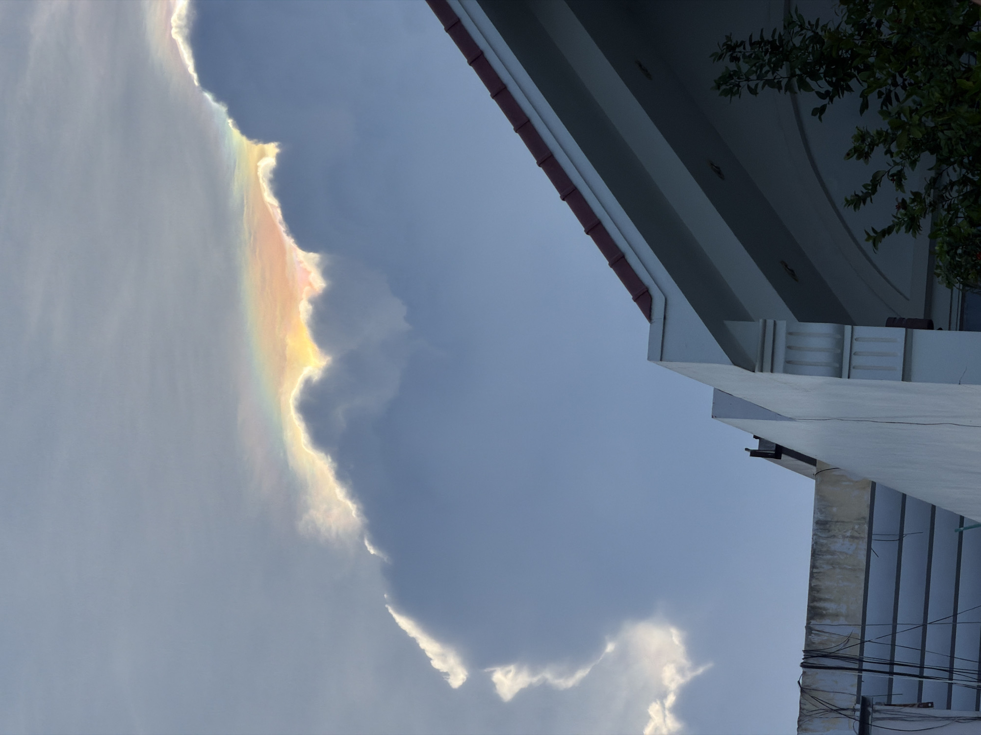 The image is taken outdoors during daytime, showcasing an iridescent cloud beside a building in Nha Trang Ward, Vietnam. The cloud exhibits a rainbow-like shimmer along its edges. The building has a multi-layered overhanging roof, with visible roof tiles. Wires hang down on one side of the building. Green foliage is present in the top right corner. The sky is blue with overcast conditions.