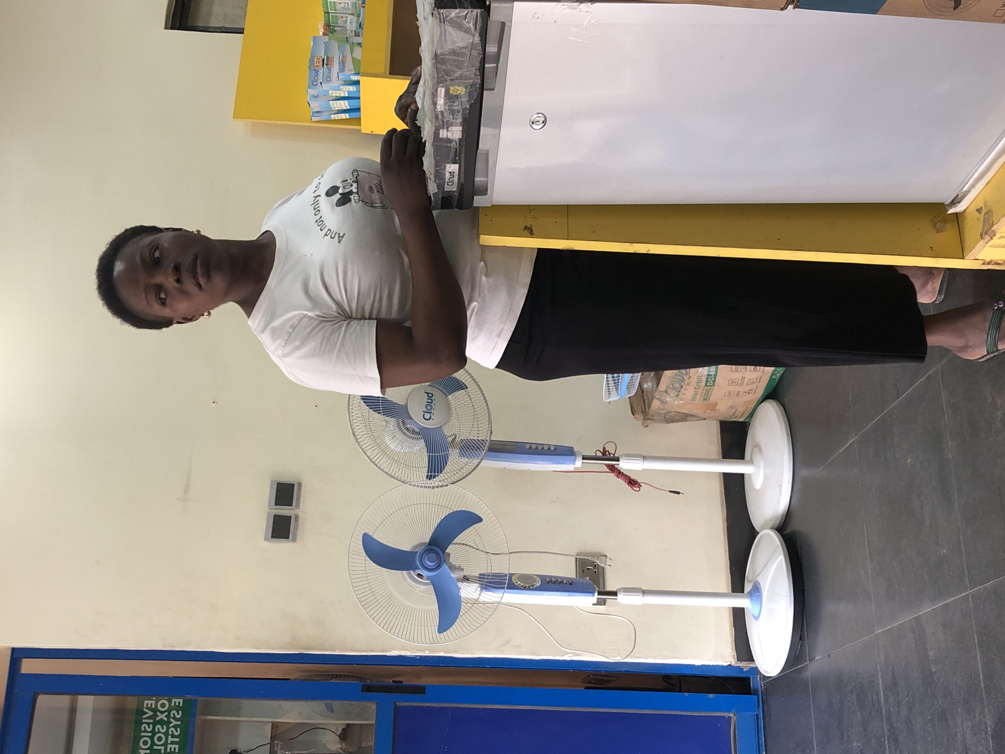 The image depicts a woman standing in a retail environment, likely a convenience store or electronics shop, in Ikeja, Nigeria. She is wearing a white t-shirt with a graphic and a black skirt. To her left, two standing electric fans are visible, one blue and white, the other blue and silver. Behind her, on the wall, are two square electrical outlets. To her right, a white refrigerator is positioned next to a yellow display shelf filled with packaged items, some labeled "Cloud LED." The flooring is dark tile. The overall impression is a snapshot of a commercial space with everyday appliances and products.
