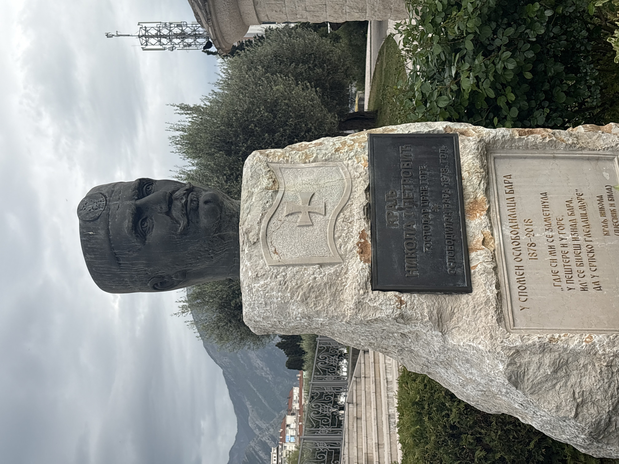 A close-up shot captures a memorial bust of King Nikola I Petrovic in Bar, Montenegro. The bust, mounted on a stone base, is surrounded by greenery, with a cityscape and a mountain visible in the background. The base includes two plaques with Cyrillic script, one indicating King Nikola's role as the liberator of Bar in 1878 and the other bearing a quote from his poetry.