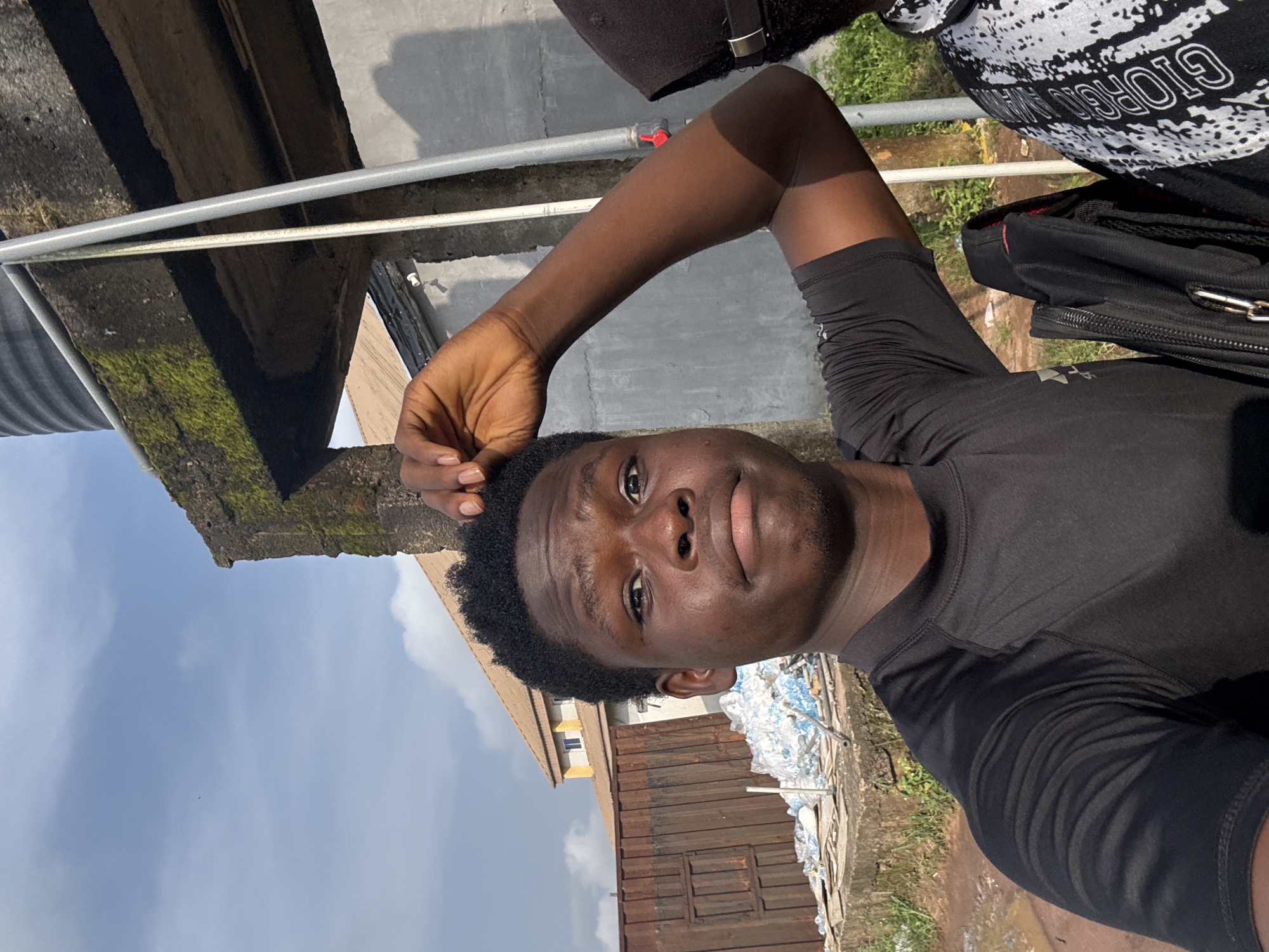 A young adult Black male is featured in a close-up, upward-angled shot, likely a selfie, taken outdoors in bright daylight in Ihiagwa, Nigeria. He has short, dark curly hair and a neutral to slightly serious expression, looking directly at the camera. He is wearing a plain black short-sleeved top, possibly athletic wear, and a black cap is visible on his head or very close by. His right arm is bent with his hand resting near his head.
The background presents a mix of urban and somewhat dilapidated structures under a clear blue sky with faint clouds. Above him, there are concrete surfaces, some showing patches of green moss, and parallel metal pipes or rails. To his right, a black bag with a zipper is visible, and behind him, a white and black patterned fabric shows the clear text "GIORGIO" followed by partial letters, likely part of "Giorgio Armani." Further back, there's a rusty corrugated metal structure, a light-colored building with visible windows, and piles of white and blue objects, possibly discarded materials or waste.