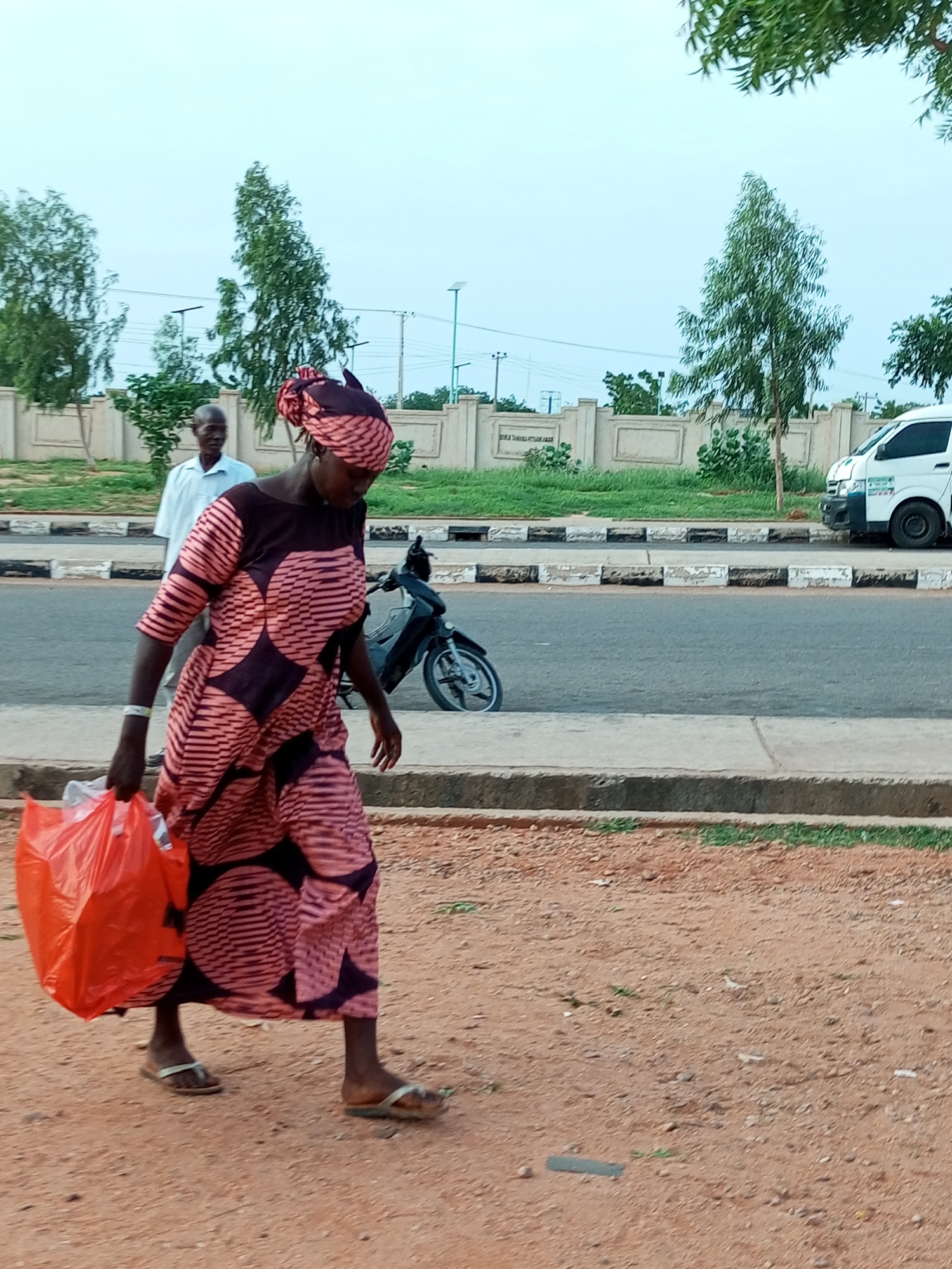 In Gombe, Nigeria, a woman walks on a dirt path parallel to a paved road. She is wearing a pink and dark patterned dress and a matching headscarf, and carrying a bright orange plastic bag in her left hand. Her feet are adorned with flip-flops. Behind her, a man in a light blue shirt stands by a beige, low wall with a metal fence on top. The road is marked with black and white stripes at its edge and carries a black motorcycle and a white van. Trees with sparse foliage dot the landscape behind the wall. The sky is overcast.