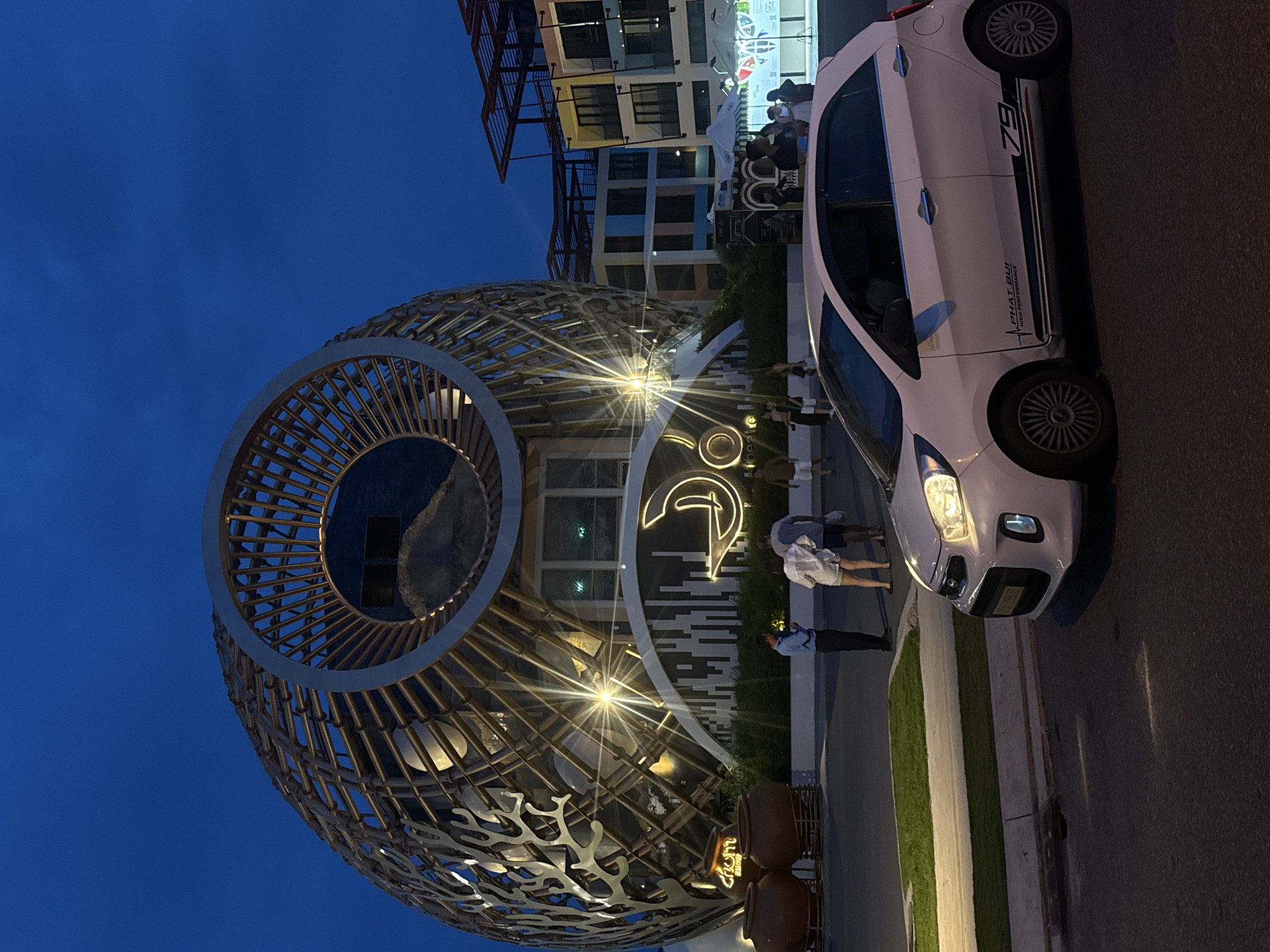 In the city of Nha Trang, Vietnam, a white Kia Picanto is parked on a dark asphalt road at dusk, its headlights illuminated. Behind the car, a large, spherical building with a distinctive, intricate wooden lattice facade stands prominently. This architectural marvel is lit from within, casting a warm glow and highlighting its unique design. In front of the building, several large, brown earthenware pots are arranged. A group of people are visible near the entrance of the building, some appearing to be staff members in uniform, while others are patrons. Further in the background, a brightly colored, multi-story building with a modern facade is visible, suggesting a commercial or entertainment district. The sky is a deep twilight blue, indicative of the evening hour.
