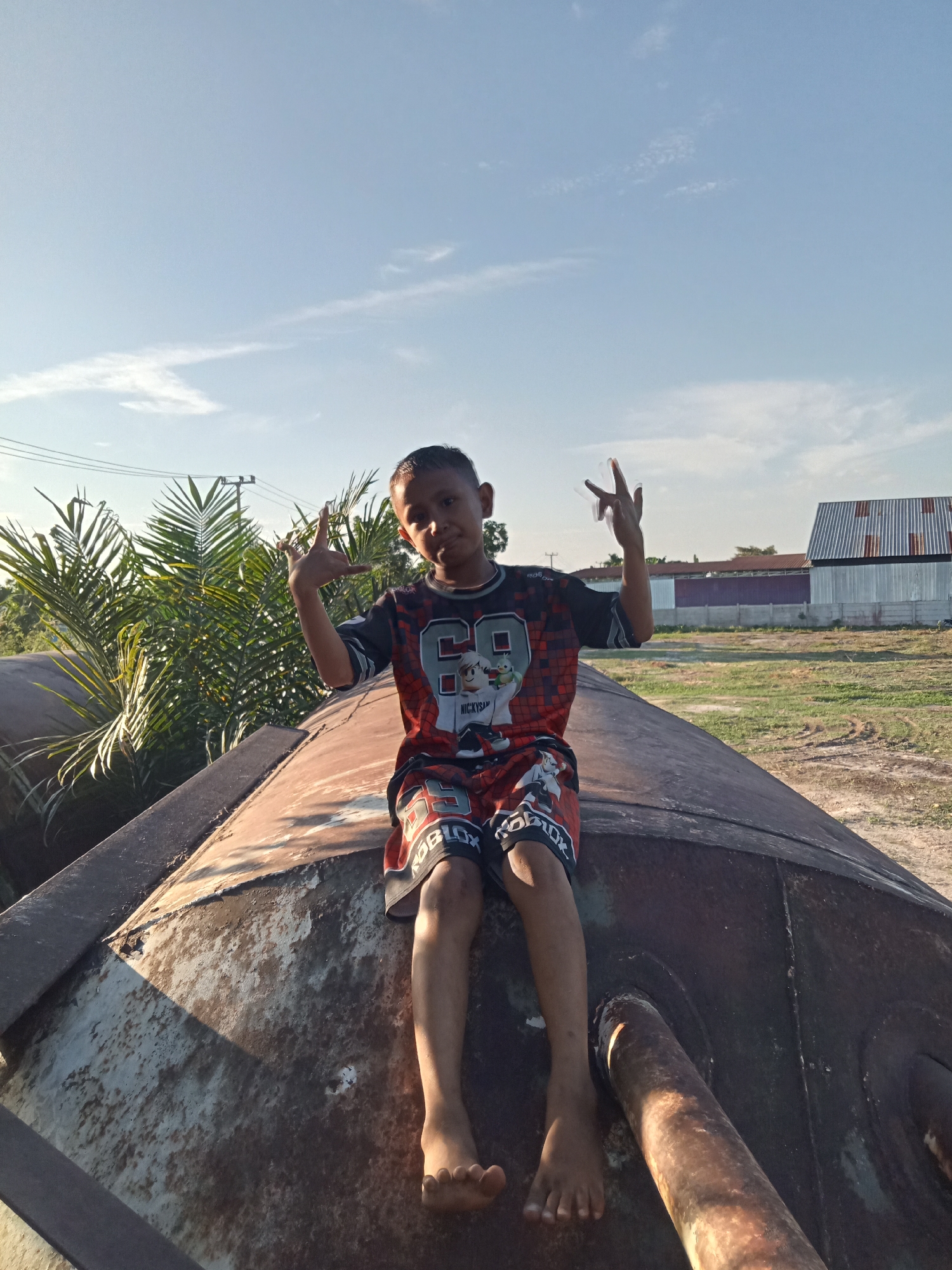 A young boy sits on a rusty, cylindrical object, possibly an old industrial tank, under a clear blue sky with scattered clouds. He is wearing a black t-shirt with red and white checkered sleeves and a graphic design featuring a cartoon character and the number "69" on the front. His shorts are black with red and white checkered trim and have a repeating "ROBLOX" pattern. The boy is barefoot and his legs are extended. He is holding his hands up, with his index and middle fingers extended in a peace sign gesture on both hands. He appears to be looking at the camera with a slight smile. Behind him, palm trees and other vegetation are visible. In the background, there are buildings with corrugated metal roofs and a grassy field. The overall scene suggests an outdoor setting, potentially in a rural or industrial area near Kualu, Indonesia. The lighting indicates it is daytime, likely late afternoon due to the angle of the sun casting shadows.