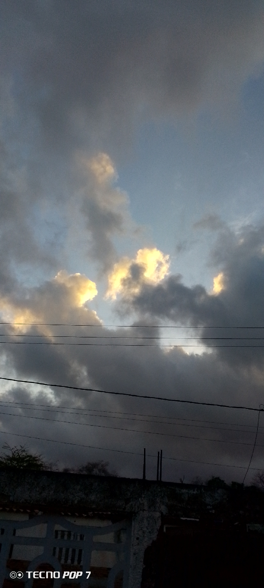 The image depicts a sky dominated by dark, ominous clouds with patches of light shining through, likely taken in Altagracia, Venezuela. Several power lines run horizontally across the middle of the frame. In the foreground, there is a glimpse of a building or structure, partially obscured in shadow. The overall environment appears to be urban, given the presence of buildings and electrical infrastructure. The image was captured with a TECNO POP 7 phone.