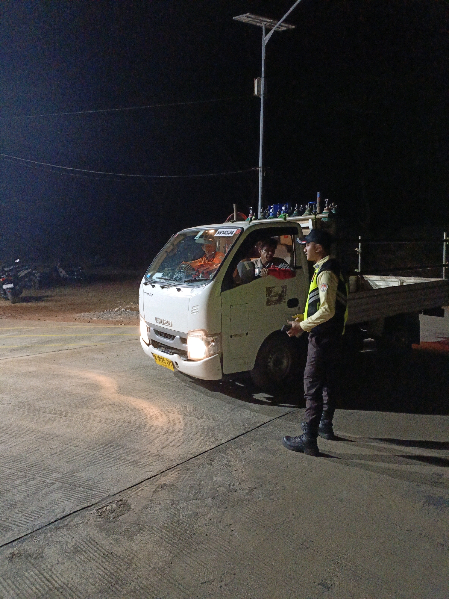 At night in Jenu, Indonesia, a white Isuzu truck is parked on a paved surface. The truck has several gas cylinders strapped to its roof. A man in an orange shirt is sitting in the driver's seat. A second man, wearing a reflective vest over a light-colored shirt and dark pants, stands outside the passenger side of the truck, facing the driver. He is holding a small device in his hand. Two motorcycles are parked in the background. A solar-powered street lamp is visible in the upper left quadrant of the image. The ground is concrete with textured markings.