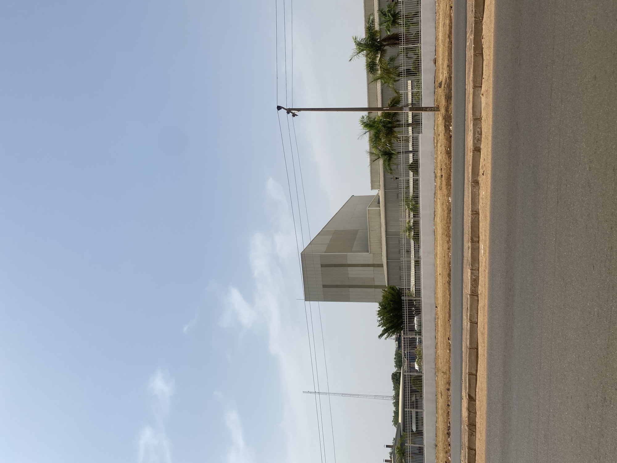 This image depicts an industrial complex in Rido, Nigeria, viewed from across an asphalt road under a clear, light blue sky with scattered white clouds. The foreground consists of a dark gray asphalt road, flanked by a light brown, sandy verge and concrete curbs. Beyond the curbs, a raised strip of dry, light brown vegetation precedes a white concrete perimeter wall topped with a light-colored metal fence.
Behind the fence, various green trees, including several palm trees, are visible within the complex grounds. A tall, dark brown utility pole with four horizontal power lines stands near the center-right. In the midground, a large, multi-section industrial building with light gray/silver metallic paneling and angular rooflines dominates, accompanied by smaller, lighter-colored buildings with corrugated roofs. To the far left, a tall, slender lattice-style communication tower rises prominently against the sky. No individuals or active vehicles are present in the scene. The environment suggests an open, industrial area under sunny conditions.