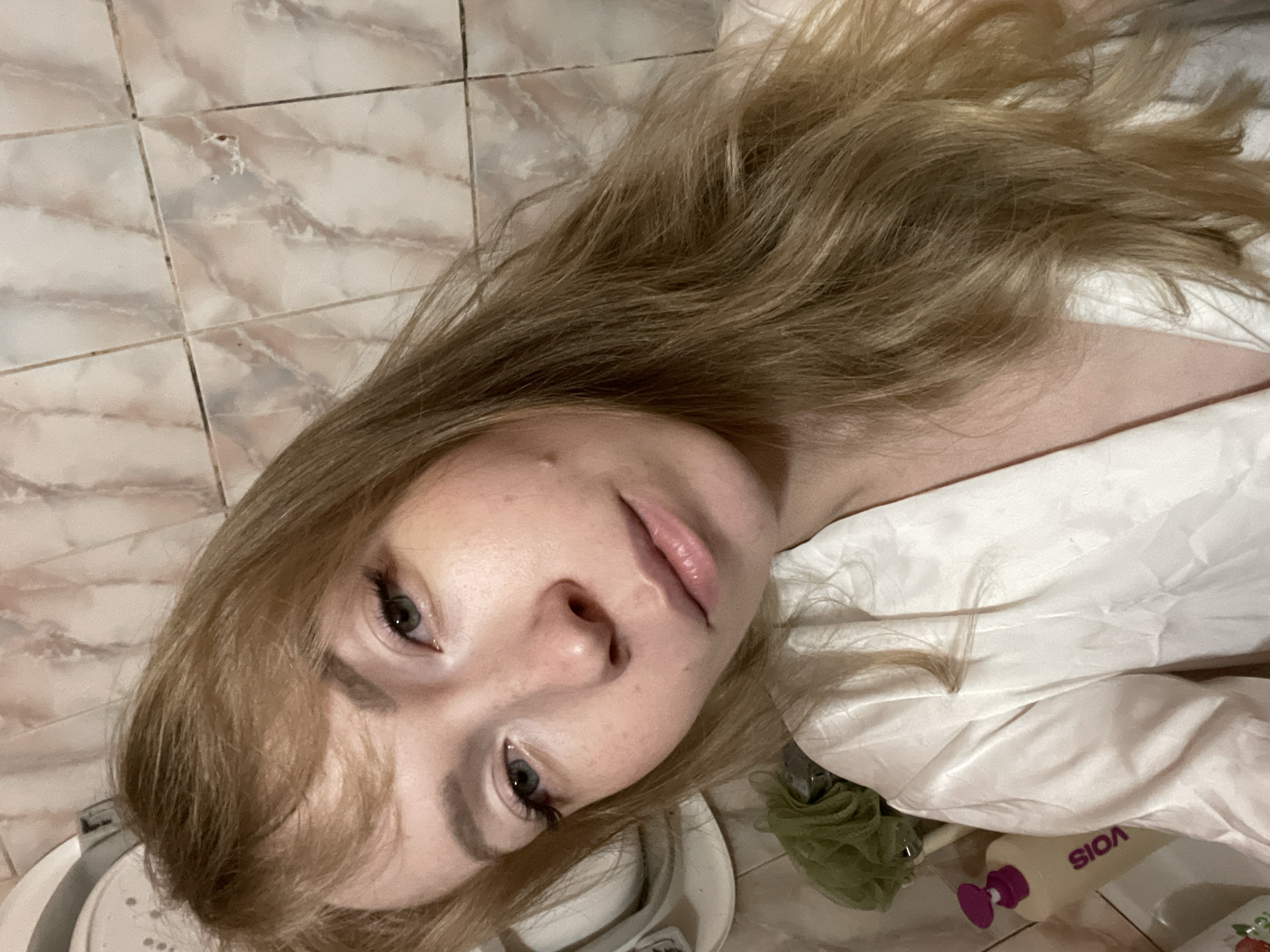 A young woman with blonde hair is lying down, facing the camera. Her head is tilted. She has fair skin, light-colored eyes, and appears to be wearing makeup. She is wearing a white garment that appears to be satin. Next to her is what appears to be a shower caddy with toiletries, including a bottle labeled "SOIA." The background is a tiled surface, which may be a bathroom or kitchen floor. The specific location in Perm, Russia, cannot be determined from the image.