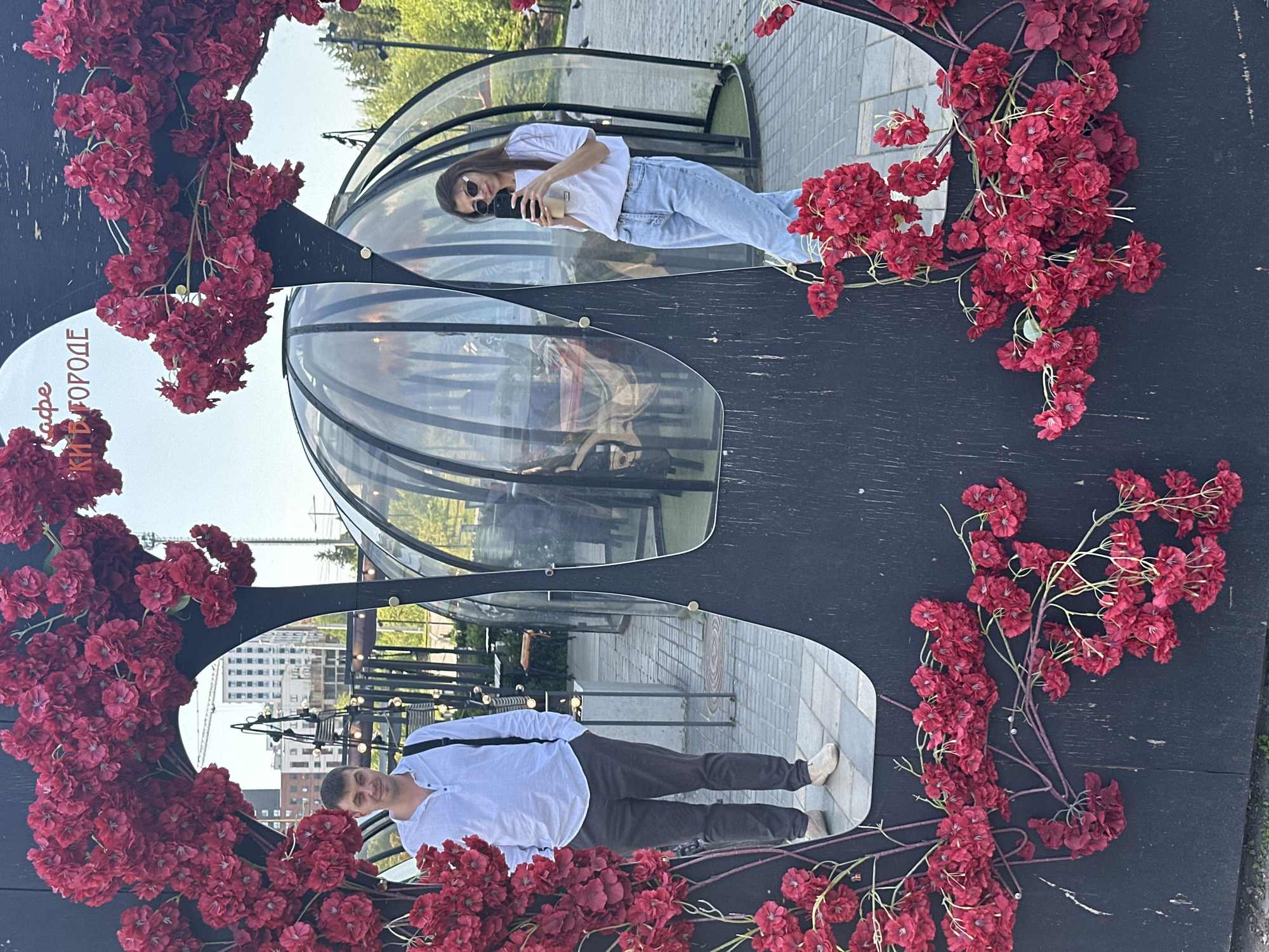A brightly-lit outdoor scene captures a uniquely designed mirror structure, adorned with vibrant red flowers. Reflected in the mirror are two distinct scenes of people and elements of the city. At the top, a woman wearing sunglasses and a white shirt and light-colored jeans is holding her phone, reflected behind a transparent dome. Below, a man in a white button-down shirt and dark pants poses for the mirror's reflection, also set against the backdrop of the city. The mirror has several reflective elements that show the city of Izhevsk, Russia including glass domes. The mirror structure is placed on a black background. The entire scene appears to be set in an urban environment, possibly a park or a public space in Izhevsk.