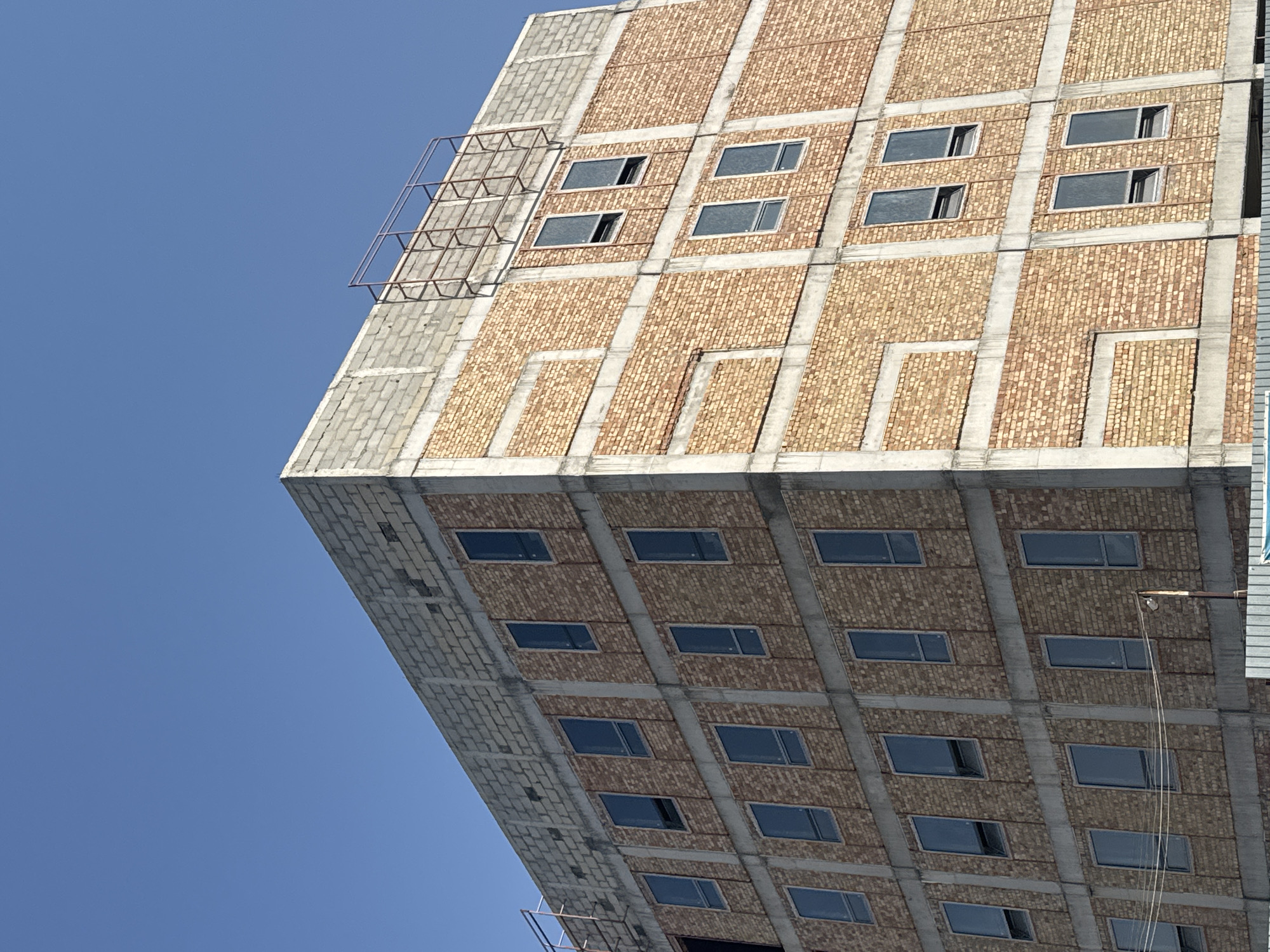 The image shows the exterior of a multi-story building under construction, possibly a residential or commercial complex, under a clear blue sky in Bishkek City, Kyrgyzstan. The building features a facade of light brown brickwork interspersed with concrete structural elements. Numerous windows, mostly dark and some with their shades partially lowered, are visible on each level. The top of the building is capped with concrete blocks, and scaffolding is present along the edge of one side. The overall impression is of a modern structure in its early stages of completion.