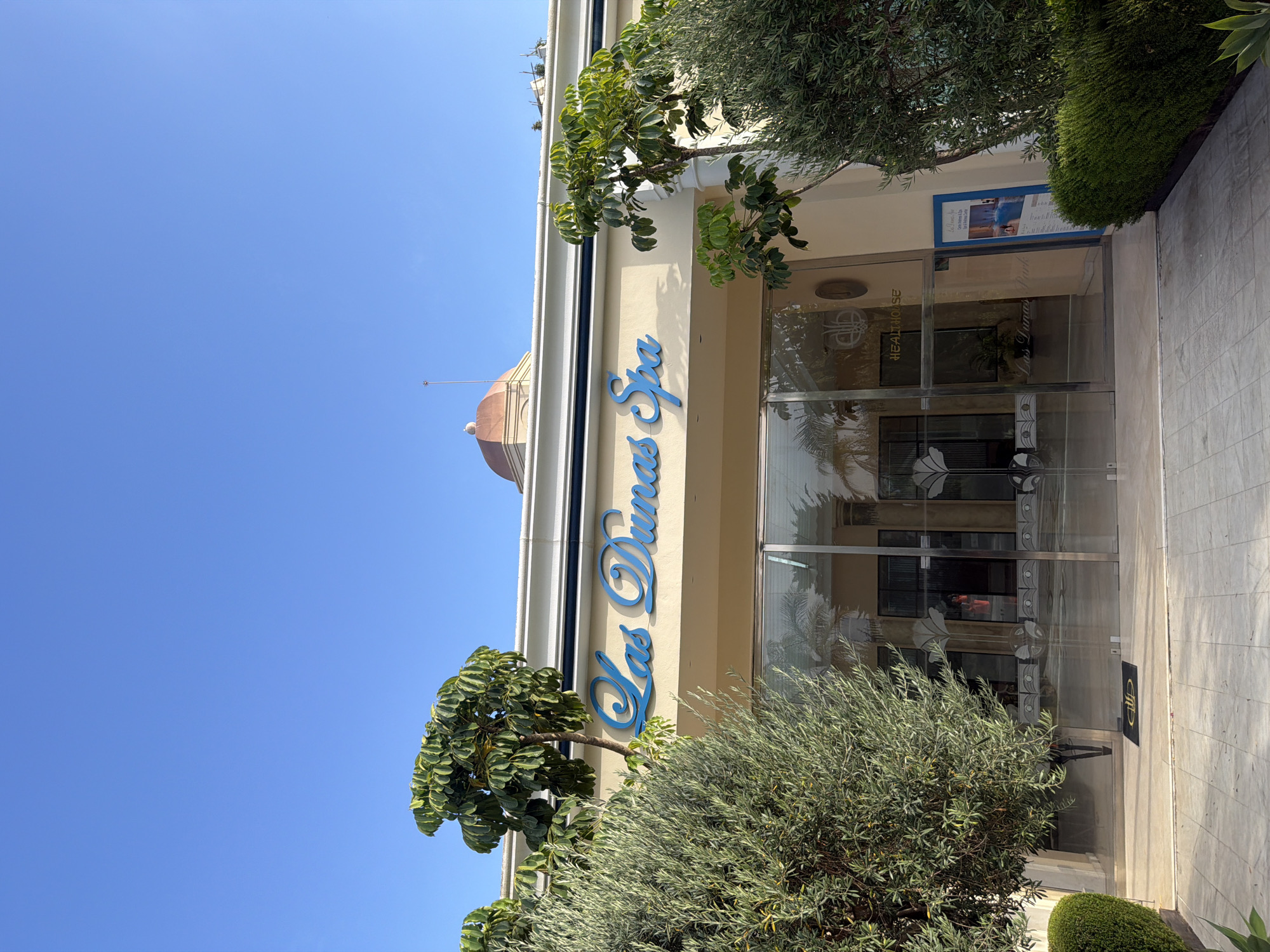 The image shows the exterior of the "Las Dunas Spa" in Estepona, Spain. The building has a light-colored facade with blue lettering spelling out "Las Dunas Spa" above a glass entrance. Green trees and bushes are visible in the foreground, flanking the entrance. The sky is clear blue. A small copper-domed structure is visible on the roof of the building. A blue information sign is placed to the right of the entrance.