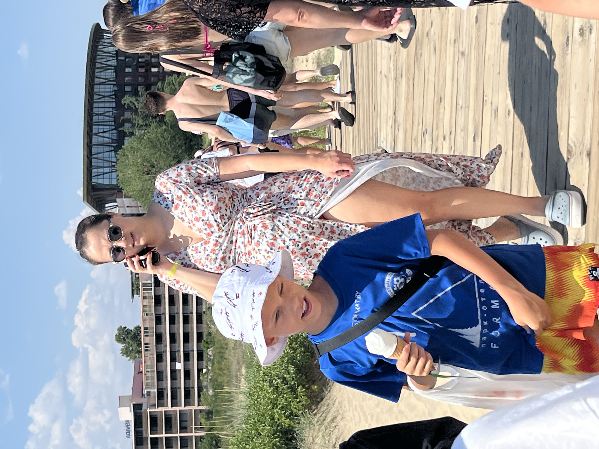 A group of people are walking on a wooden boardwalk on a sunny day. In the foreground, a young boy wearing a blue t-shirt, a white bucket hat, and patterned shorts is holding an ice cream cone. To his right, a woman in a floral wrap dress is talking on her phone. In the background, several other individuals are walking along the boardwalk, some carrying bags. Beyond the boardwalk, there are sandy areas with vegetation and buildings, including a large hotel-like structure and a distinctive modern building with a curved roof. The sky is blue with scattered clouds. The location is identified as Nesebar, Bulgaria.