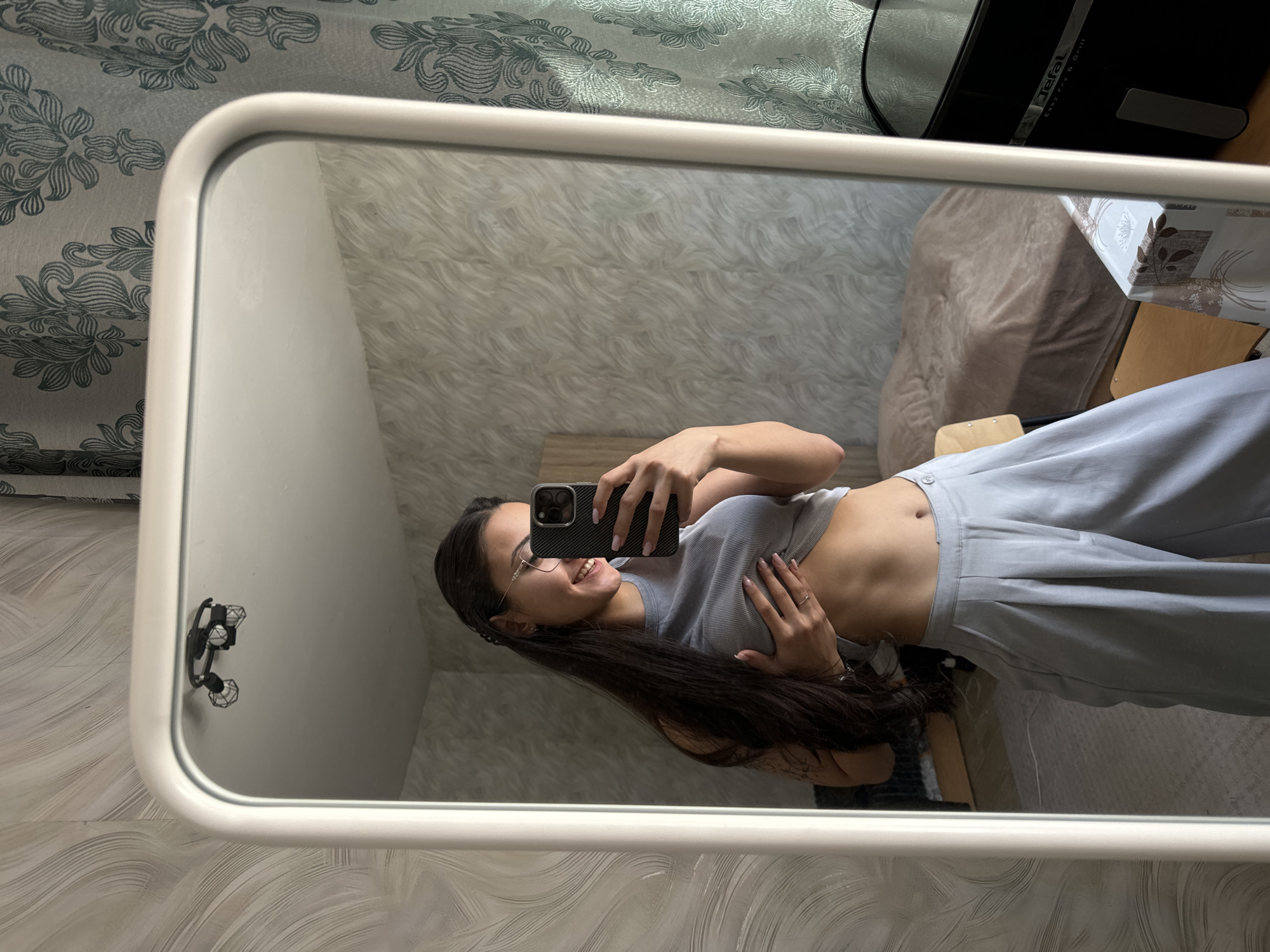 A young woman is captured in a mirror selfie, smiling broadly with her head tilted to the side. She has long, dark hair and wears glasses, a light grey ribbed crop top that reveals her midriff, and matching light blue or grey wide-leg trousers. She holds a black smartphone in her left hand to take the picture, while her right hand rests on her abdomen. A ring is visible on one of her fingers.
The mirror has a white or light-colored frame. The reflection shows an indoor setting, likely a room in a home in Sofia, Bulgaria. The background features a wall with patterned wallpaper, displaying swirling or floral motifs in grey and light blue tones. Above this, a section of a plain, light-colored wall is visible. Within the mirror's reflection, behind the woman, there is a light-colored upholstered sofa or piece of furniture and a table covered with a patterned tablecloth. A black electronic device is also visible, bearing the text "Tefal" and "Easy Fry & Grill." To the left, a black wall-mounted fixture with two decorative diamond-shaped elements is attached to the wall. The lighting suggests it is daytime, and the woman appears to be happy.
