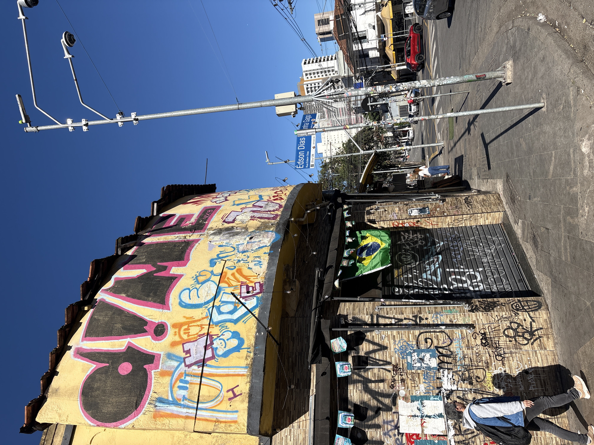 The image depicts a street scene in São Paulo, Brazil, characterized by vibrant graffiti on a yellow building facade and a brick wall. A person, viewed from behind, walks on a concrete sidewalk, carrying a backpack. The building displays large graffiti with black and pink lettering against a yellow background, and smaller tags and drawings on the brickwork. A Brazilian flag hangs from the building. Street signs indicate "Edson Dias" and "Rua Edson Dias," suggesting the location is a street named Edson Dias. A street light with multiple arms and cameras stands prominently on the sidewalk. The street is paved, with cars, including a red vehicle, visible in the background. The sky is clear and blue. The architecture includes multi-story buildings in the distance.