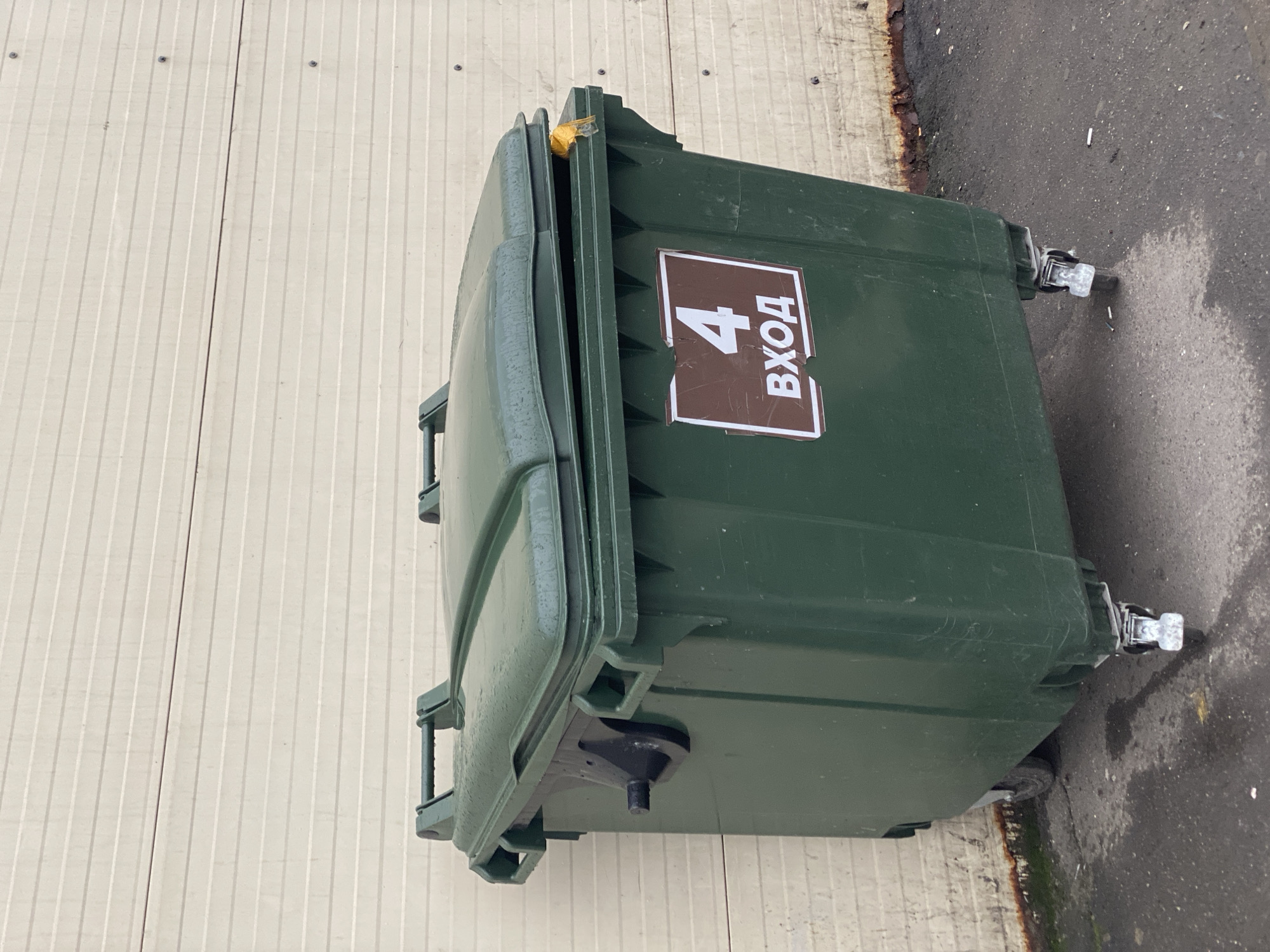 A large, dark green dumpster with a partially open lid stands against a light-colored, horizontally ribbed wall. The dumpster has two handles on the lid and appears to be made of plastic. A sign with a white border and a brown background is affixed to the front of the dumpster. The sign displays a large white number "4" above the white Cyrillic text "ВХОД" (VHOD), meaning "entrance". A black fixture with a protruding cylindrical part is attached to the left side of the dumpster. The dumpster is on wheels, with at least two visible casters, one of which appears to be locked. The ground in front of the dumpster is a dark, wet asphalt surface with some debris, including cigarette butts. A faint rusty stain runs along the bottom of the wall. The scene is outdoors.