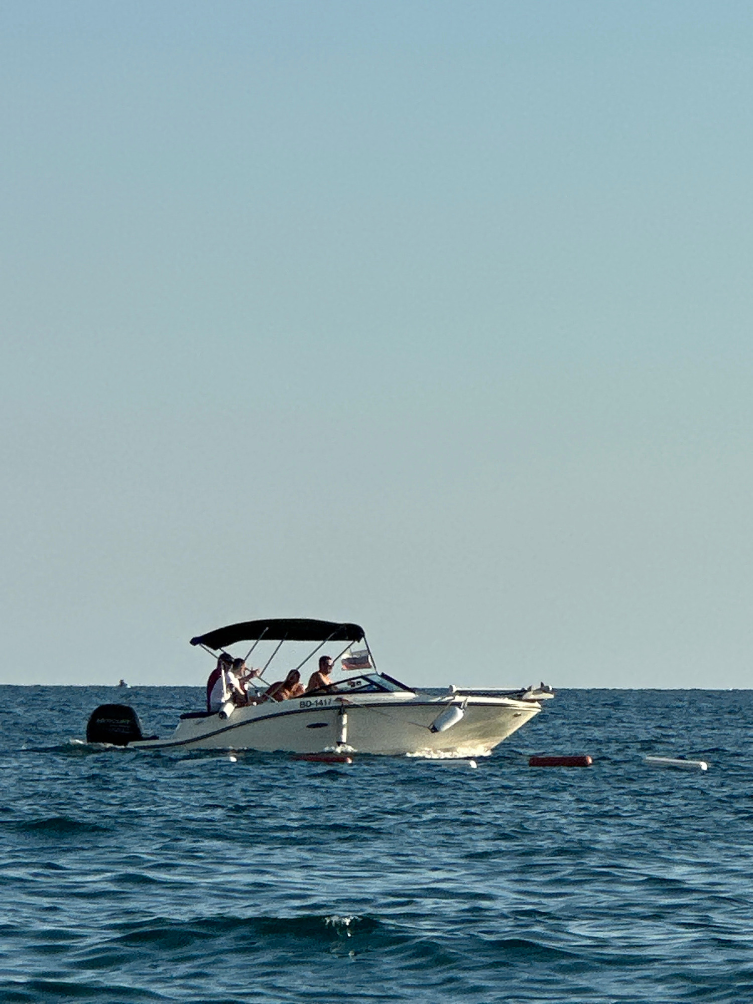 A speed boat with four people on board navigates the blue waters of the Adriatic Sea, near the coast of Boreti, Montenegro. The boat, white with a black canopy, has a Russian flag at the stern and the license plate BD-1417. The people are casually dressed, some standing and looking around, others seated. The sea is calm with moderate waves, and the sky is clear and blue. Several orange and white buoys are visible in the water around the boat. The overall scene depicts a recreational boat trip on a sunny day.