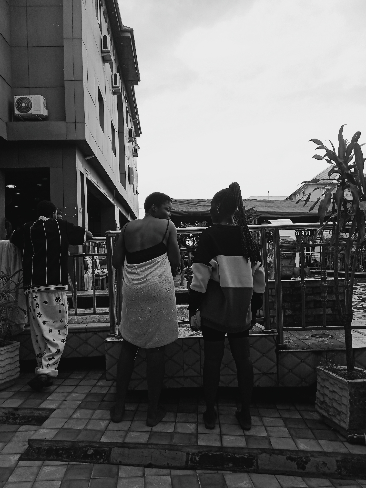 The image, in black and white, shows three individuals standing near a railing overlooking a pool or water feature in Onitsha, Nigeria. A man is visible on the left, wearing a vertically striped top and pants patterned with paw prints. A woman stands to the right of him, wrapped in a towel. Another woman is positioned further to the right. She's wearing a color-blocked sweatshirt and leggings. The location is defined by the architectural style of the building behind them, featuring an external air conditioning unit, the plants in pots, and the overall setting, all suggestive of an urban environment in Onitsha. The background includes additional structures and foliage.