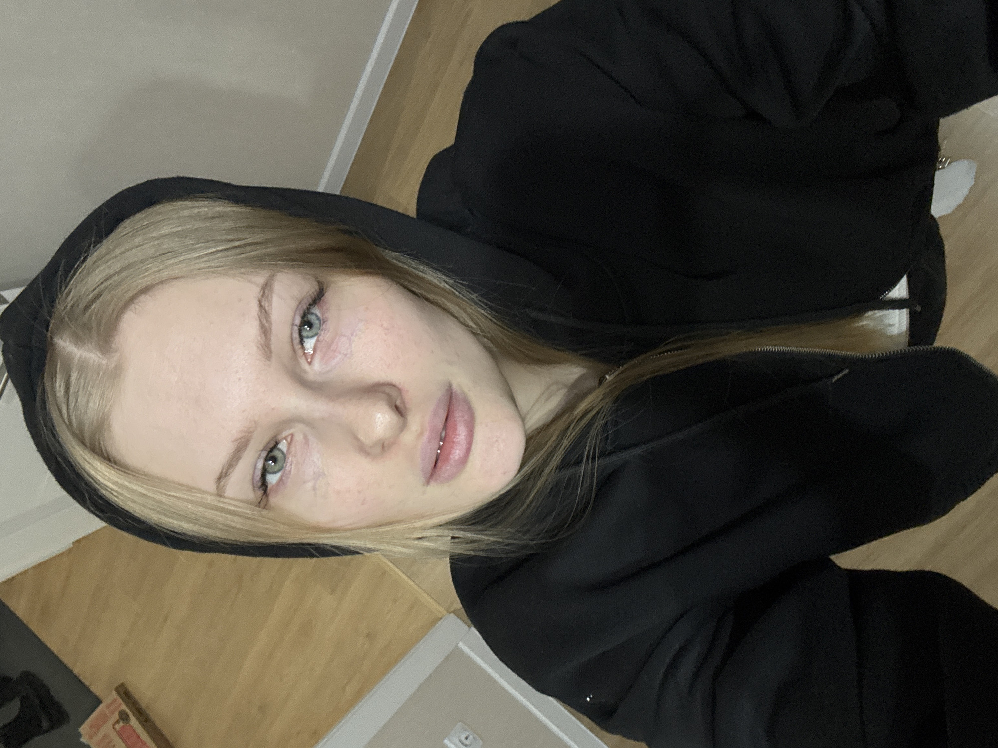 A rotated portrait-style photo features a young woman with blonde hair, fair skin, and light blue-green eyes. She is positioned centrally, looking directly at the camera with a neutral, possibly pensive or tired, expression. Her eyes exhibit some visible redness around the whites and lower lid. She is wearing a black hooded sweatshirt or jacket, with the hood pulled up to frame her face. Natural skin texture, including some minor blemishes, is visible. A small, shiny labret piercing is discernable on her lower lip. Her lips are naturally colored and slightly parted.
The setting appears to be indoors, indicated by a light-colored wooden floor, possibly laminate or parquet, and plain off-white or light grey walls in the background. Some indistinct objects, including a white, textured item that might be a rug or blanket, are partially visible on the floor in the lower portion of the image. The lighting is soft and ambient, consistent with indoor conditions, and does not suggest a specific time of day. The overall mood conveyed by the subject's expression and the simple setting is quiet and understated. There is no visible text.
