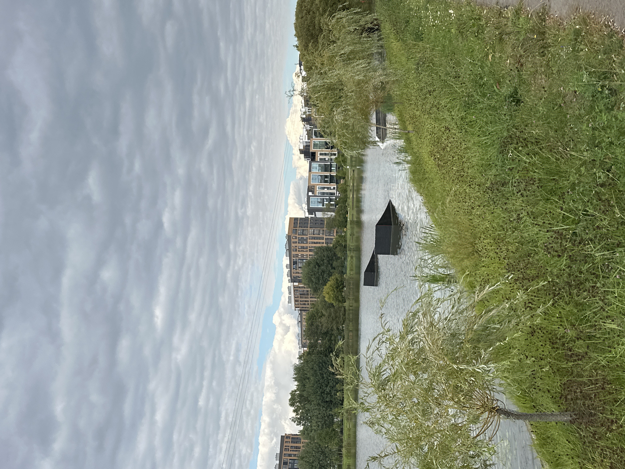 This is a full-shot, vertical, eye-level, outdoor, daytime photograph of a body of water with structures and buildings in the background, located in Minsk, Belarus. The foreground features a path on the right, lined with tall, green grass and a small willow tree with cascading branches. The water occupies the middle ground and is calm, with two dark, angular structures, possibly ramps for watersports, partially submerged. In the background, across the water, there is a dense line of trees separating the water from a row of modern, multi-story buildings with balconies and large windows. The sky above is overcast with scattered clouds.