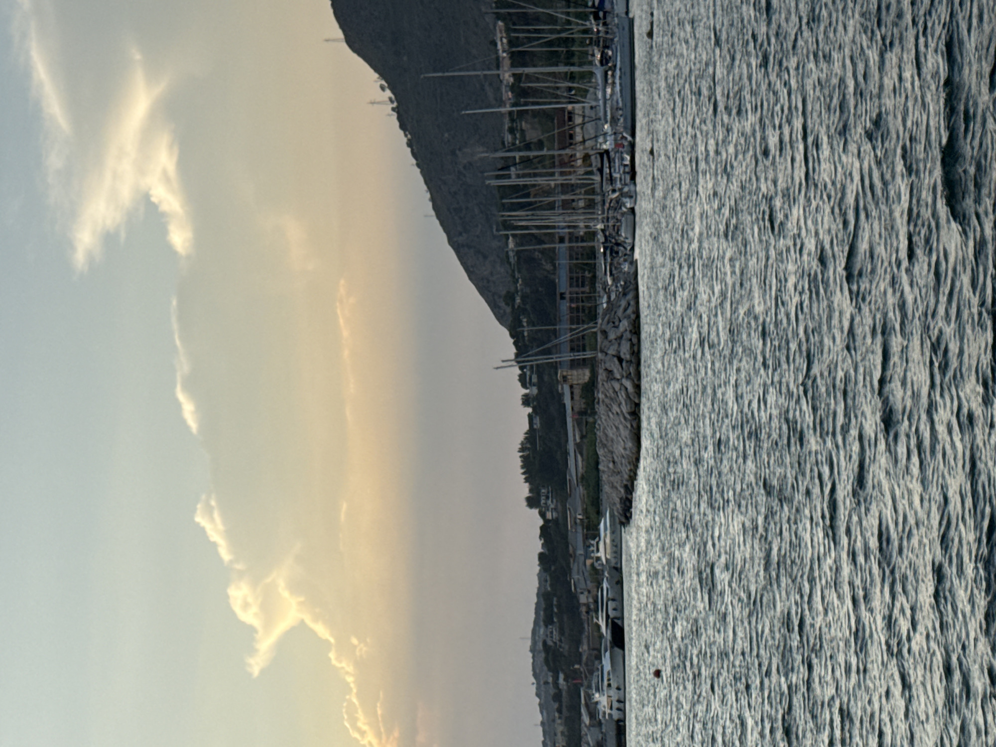 This is a photograph of the port of Pozzuoli, Italy, taken from the water. The image captures a scenic view of yachts and sailboats docked in the marina, with a mountainous backdrop. The sky is partly cloudy, with the sun setting and casting a warm glow on the clouds.
In the foreground, the water of the bay is choppy, with small waves creating a textured surface. A red buoy is visible floating in the water to the left of the frame.
The middle ground features the marina, with several white yachts moored along a stone breakwater. Beyond the yachts, there are numerous sailboats with their masts rising high into the air. The breakwater is constructed from large, rough stones.
In the background, a lush green hillside rises steeply, dotted with trees and some buildings. The hillside is crowned with what appear to be electrical pylons. The setting sun creates a soft, diffused light, giving the scene a serene and peaceful atmosphere.