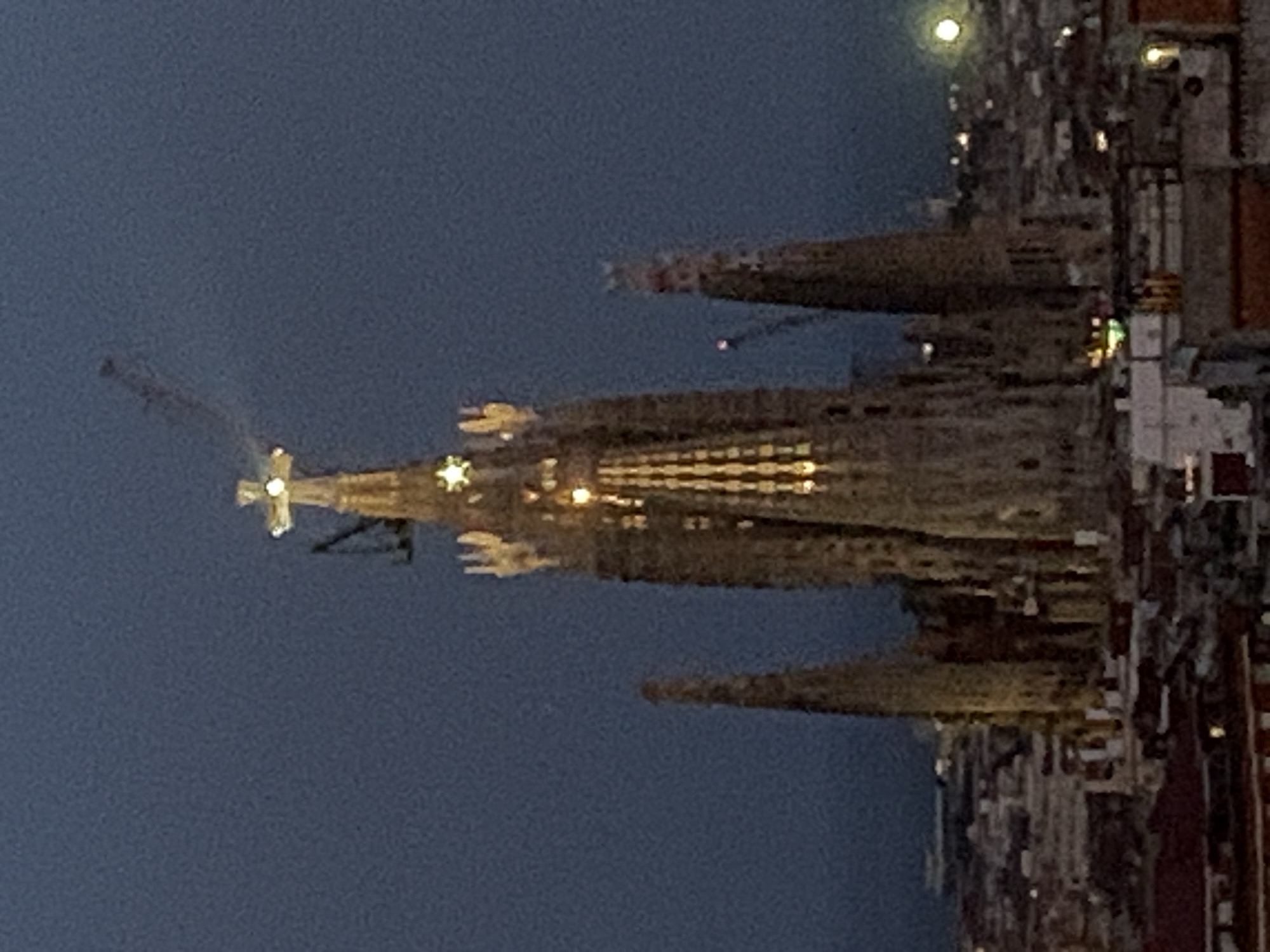 The image depicts the Sagrada Familia basilica in Barcelona, Spain, at dusk or night. The basilica's towering spires are illuminated, highlighting its intricate Gothic and Art Nouveau architecture. A large cross tops the central spire, and smaller spires rise on either side. The sky is a deep, dark blue, typical of twilight. In the background, the city of Barcelona is visible, with numerous lights twinkling across the urban landscape, suggesting a densely populated area. A crane is visible near the top of one of the spires, indicating ongoing construction or renovation work on the basilica. The overall impression is one of a grand, iconic structure set against a darkening sky over a vibrant city.