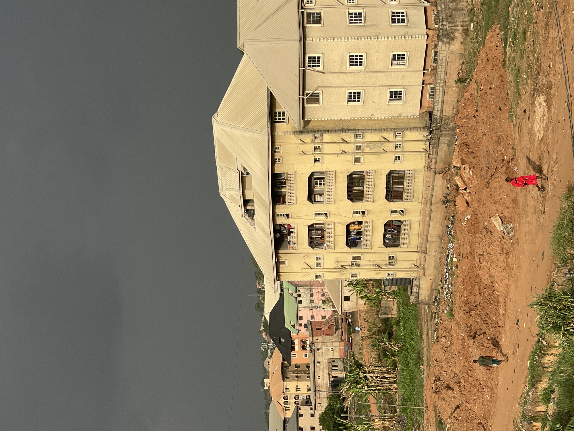 The image shows a view of an urban scene in Onitsha, Nigeria. A tall, multi-story building with a light-colored facade dominates the right side of the frame. Clothes are hanging on some of the balconies. To the right of the building, a dirt path runs along its edge. Two people are walking on the path, one wearing red clothing and the other wearing green clothing. Beyond the building, there are other buildings of various colors and sizes, with a mix of residential and commercial architecture. The sky is dark and overcast, suggesting a storm is approaching. The environment appears to be a mix of undeveloped land and built-up urban areas.