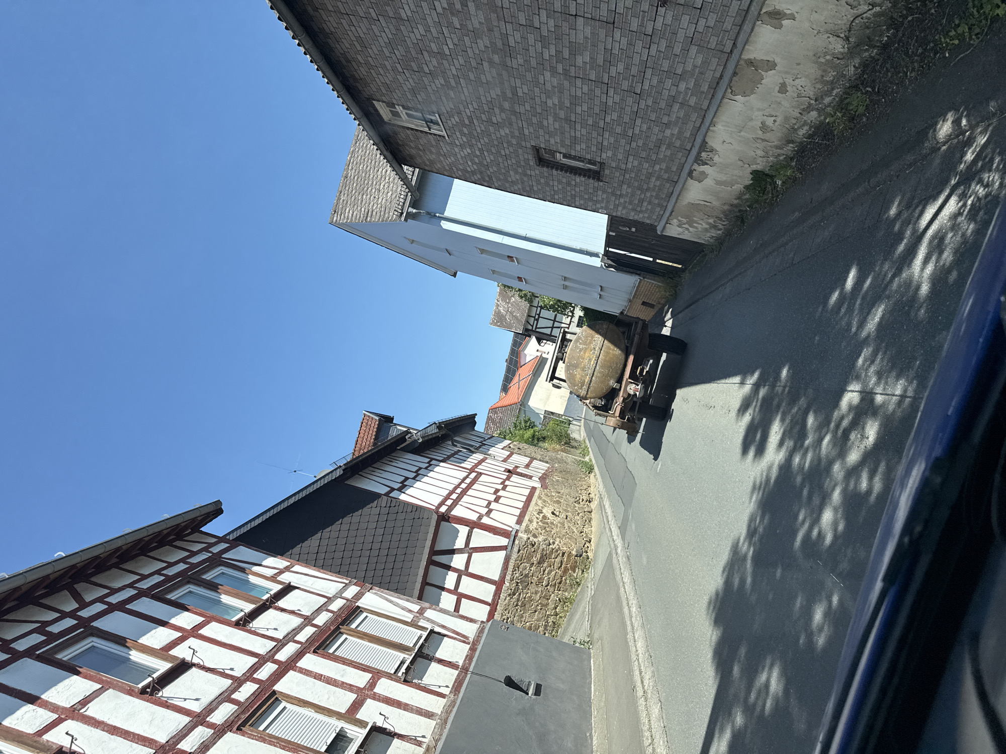 A paved road runs through a European village under a clear blue sky. On the left, a traditional half-timbered building with white stucco and red-brown beams is visible. The building has multiple windows with white shutters. A stone foundation supports the lower portion of the house. On the right, a light blue building with several windows stands next to a grey brick building with a textured facade and a weathered section of plaster at its base. Further down the road, another half-timbered building with a red-tiled roof is partially visible. In the middle of the road, a rustic, old trailer with a large, round, metallic tank is being towed. The trailer has two wheels on each side and appears to be moving away from the viewer. Shadows of trees are cast across the road and the side of the blue building. The perspective suggests the image was taken from inside a vehicle. The scene is characteristic of a rural or historic town in Germany, specifically Volpertshausen, given the architectural style.