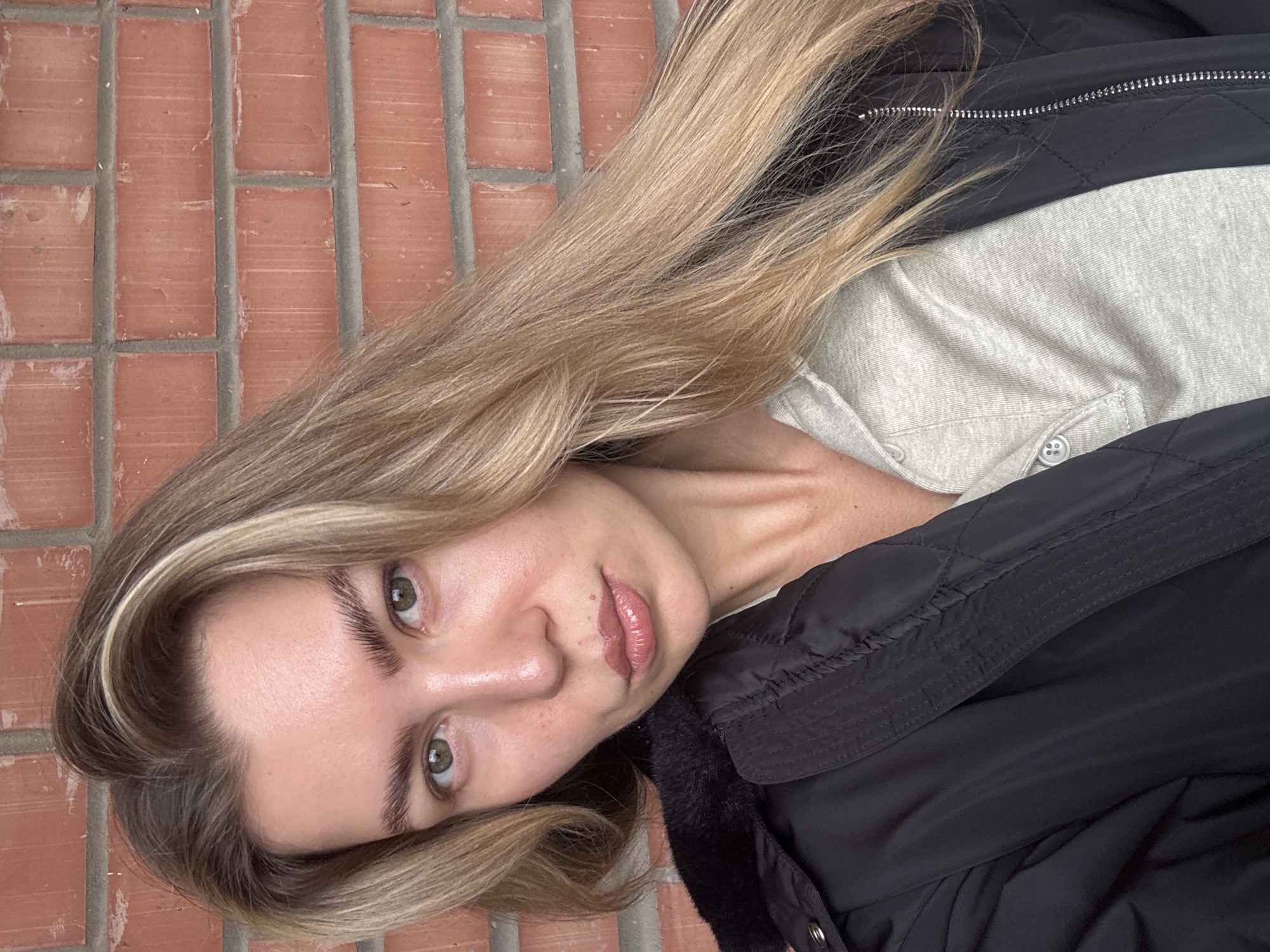 A medium shot photograph shows a young white woman with fair skin, light green eyes, and long, wavy blonde hair. She is wearing a black puffer jacket with a fur-lined hood over a light grey Henley shirt. The woman is looking at the camera with a neutral expression. Her lips are slightly parted, and she has a mole on her upper lip. Her eyebrows are dark and full. The background is a red brick wall. The lighting is soft and natural. The image appears to have been taken outdoors.