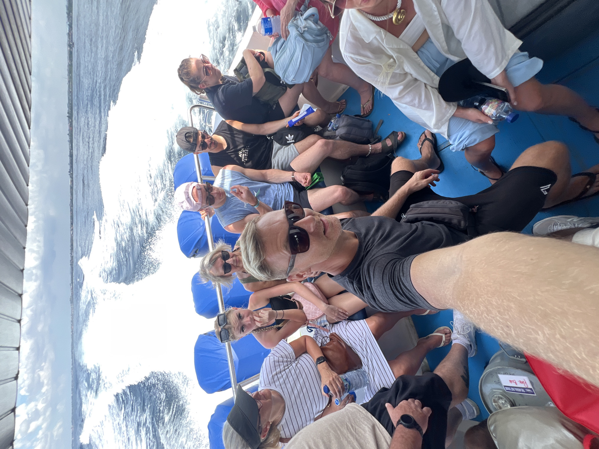 A group of approximately ten individuals is depicted on a boat, presumably on a tour or journey through open water, suggested to be in Indonesia. The image is a selfie taken by a man in the foreground on the right, wearing dark sunglasses and a black t-shirt, who is partially visible. The boat appears to be moving at speed, generating a significant white wake in the water on the left side of the frame.
The passengers are casually dressed in shorts, t-shirts, and light blouses, suitable for warm weather. Many are wearing sunglasses and some have hats or caps. They are seated on blue cushioned benches, with a blue deck visible beneath their feet. Various personal items are scattered around, including several plastic water bottles (one clearly showing "AQUA" and "PURE LIFE" labels), backpacks, and fanny packs (one with an "Adidas" logo visible).
The scene is set during the daytime under what appears to be bright, clear skies. In the distance, a coastline with faint structures or vegetation can be seen. The general atmosphere suggests leisure and enjoyment of the boat ride, with some individuals smiling or looking relaxed. In the lower right corner, a grey container-like object has "PURA VIDA" written on it.