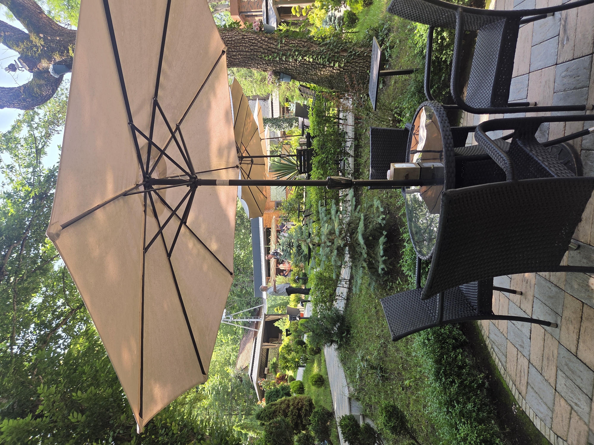 In the city of Dilijan, Armenia, an outdoor seating area is depicted. A large, tan patio umbrella shades a round, glass-top table with four dark wicker chairs surrounding it. The table has a napkin dispenser and a small umbrella. Behind this seating arrangement, a pathway paved with rectangular stones leads through a lush garden filled with various green shrubs and trees. In the background, there are several outdoor structures, including a covered gazebo with a brown tiled roof and a longer, modern building with glass windows. A few individuals are visible in the background, some standing and interacting near the buildings. The overall environment is natural and serene, with abundant greenery and clear daylight.