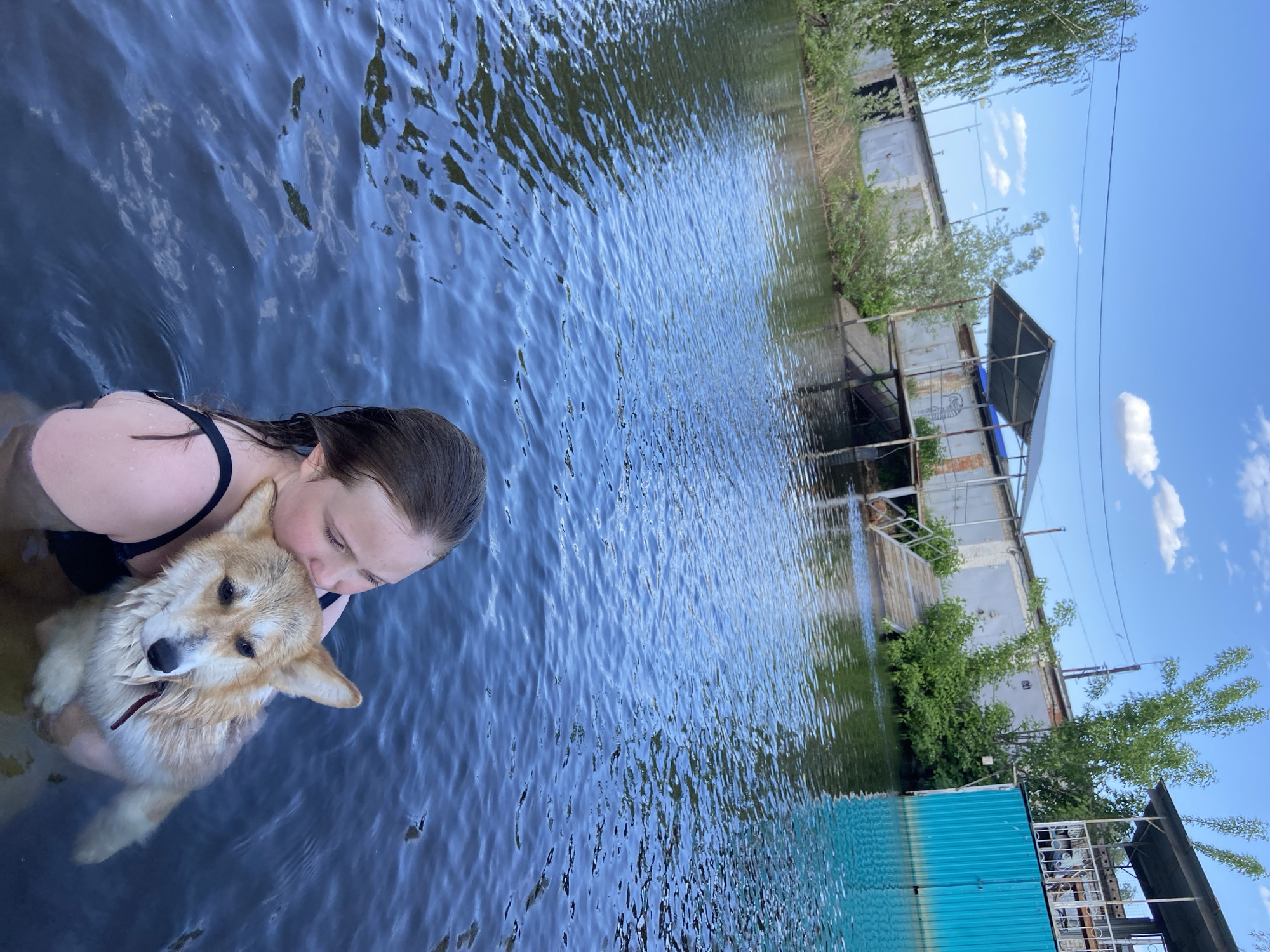 A woman and a corgi dog are in the water of a body of water in Chaikovsky, Russia. The woman is wearing a black top and has dark hair. She is holding the corgi close to her face. The dog is wet and has light brown and white fur. The water is dark and rippled, and the background includes buildings and vegetation.