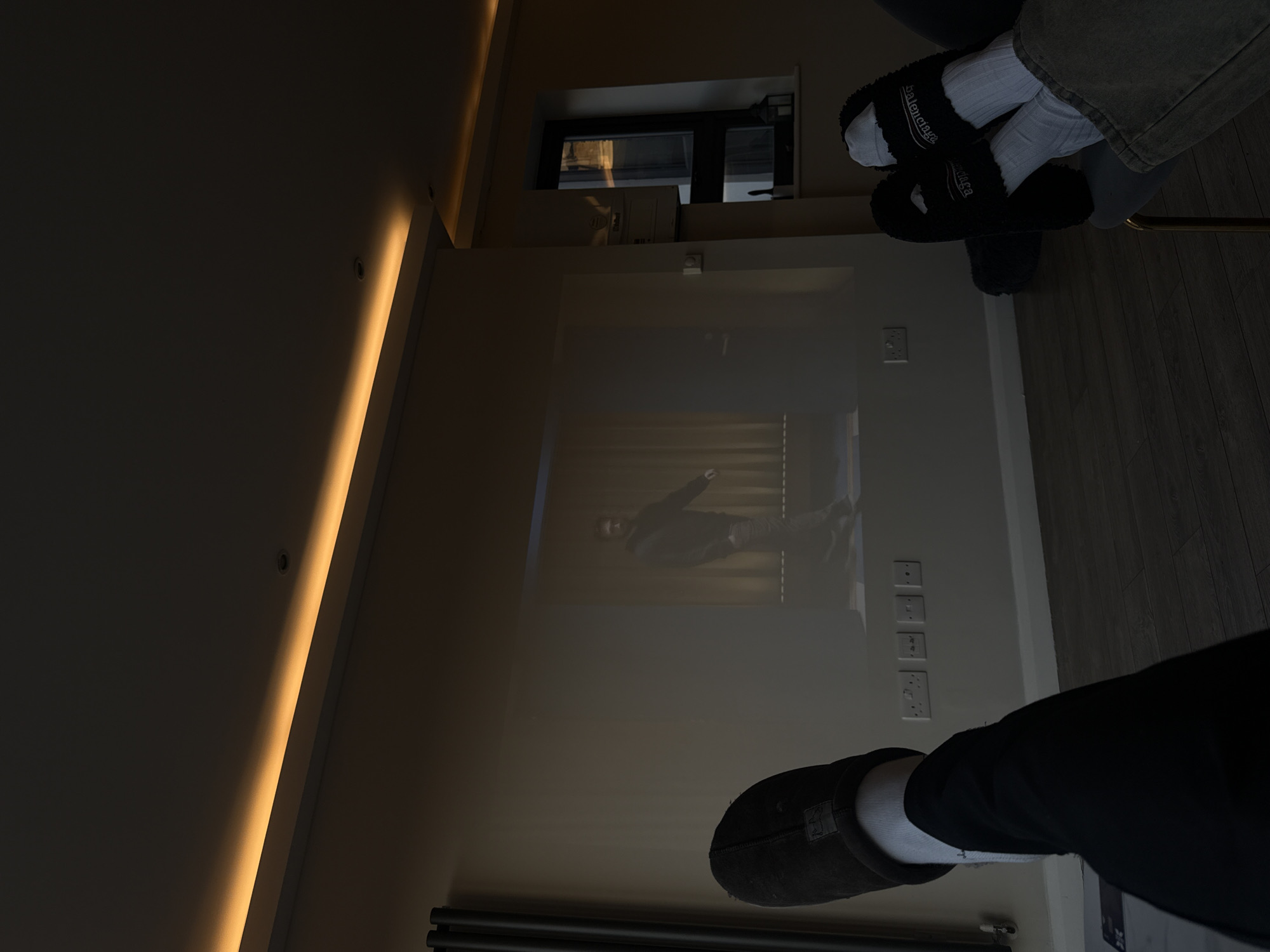 The image depicts an indoor scene in London, United Kingdom, characterized by dim ambient lighting and a projected display. Two individuals are present, indicated by their lower extremities.
On the left, a person's right foot, clad in a dark brown UGG-style slipper over a white sock and dark trousers, is visible in the foreground, elevated. On the right, another person's feet, wearing black fluffy Balenciaga slides over white socks and light-colored trousers, are elevated and resting on a dark grey chair with gold-colored legs.
A large, rectangular image is projected onto a light-colored wall, serving as the primary focal point. The projection displays a male figure with a beard, wearing a dark hoodie and dark trousers, in a walking or standing pose against a background featuring vertical, curtain-like elements.
The room's ceiling features warm orange LED strip lighting along its perimeter and at least one recessed spotlight. Below the projected image on the wall, three white Type G electrical sockets and one white switch are mounted. To the far left, a dark, vertical panel radiator is partially visible. To the right of the projection, a dark-framed vertical window looks out onto an obscured exterior view, possibly reflecting another building. Above this window, a white wall-mounted unit, possibly a Vaillant boiler, is affixed. The floor is covered with a dark wood-effect laminate. A dark, lantern-style decorative object is present on the windowsill beneath the window.